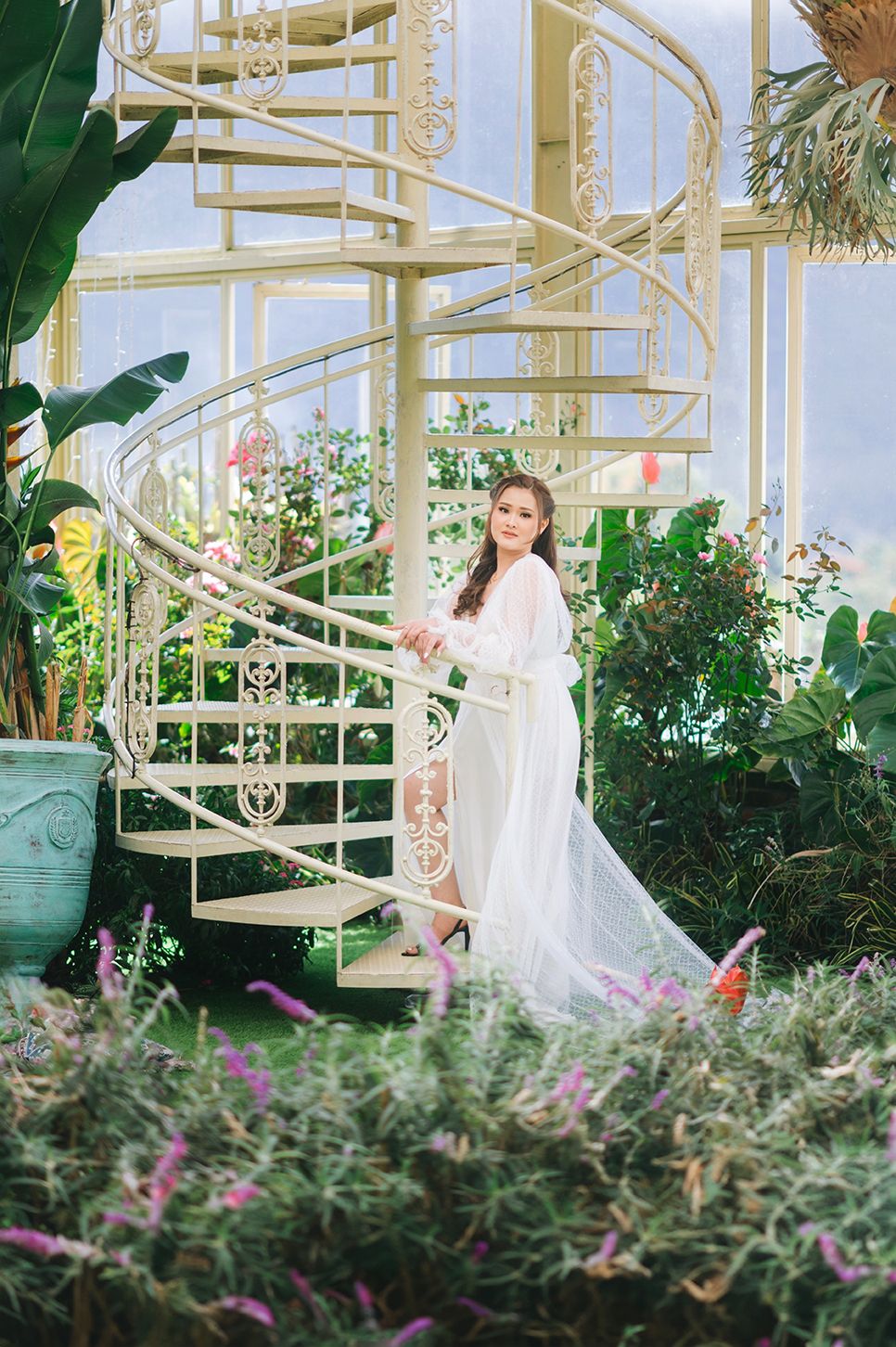 Woman in white gown poses on ornate spiral staircase surrounded by lush garden greenery.