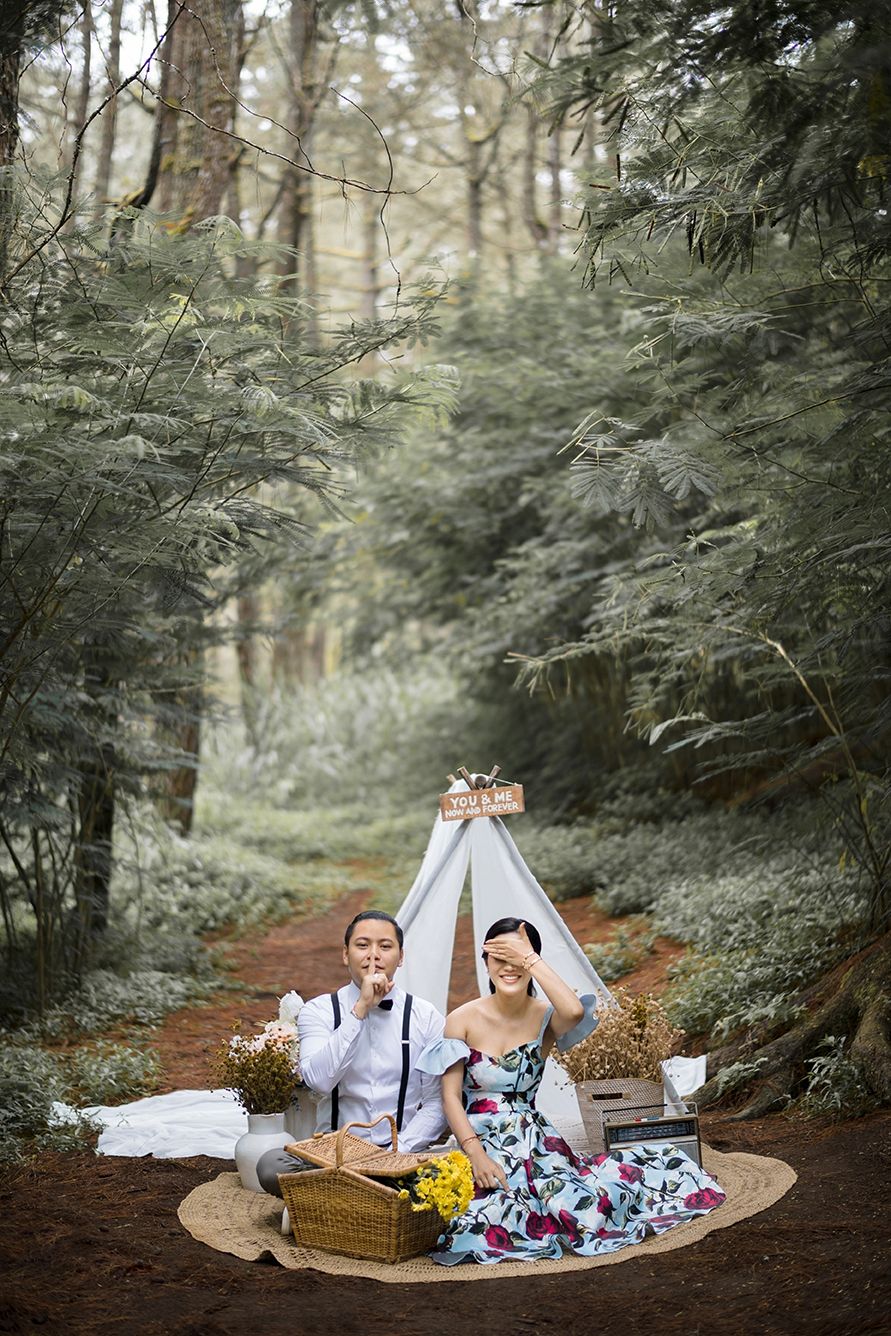 Couple in a forest picnic setting with a white teepee, floral dress, yellow flowers, and a wicker basket on a round mat.