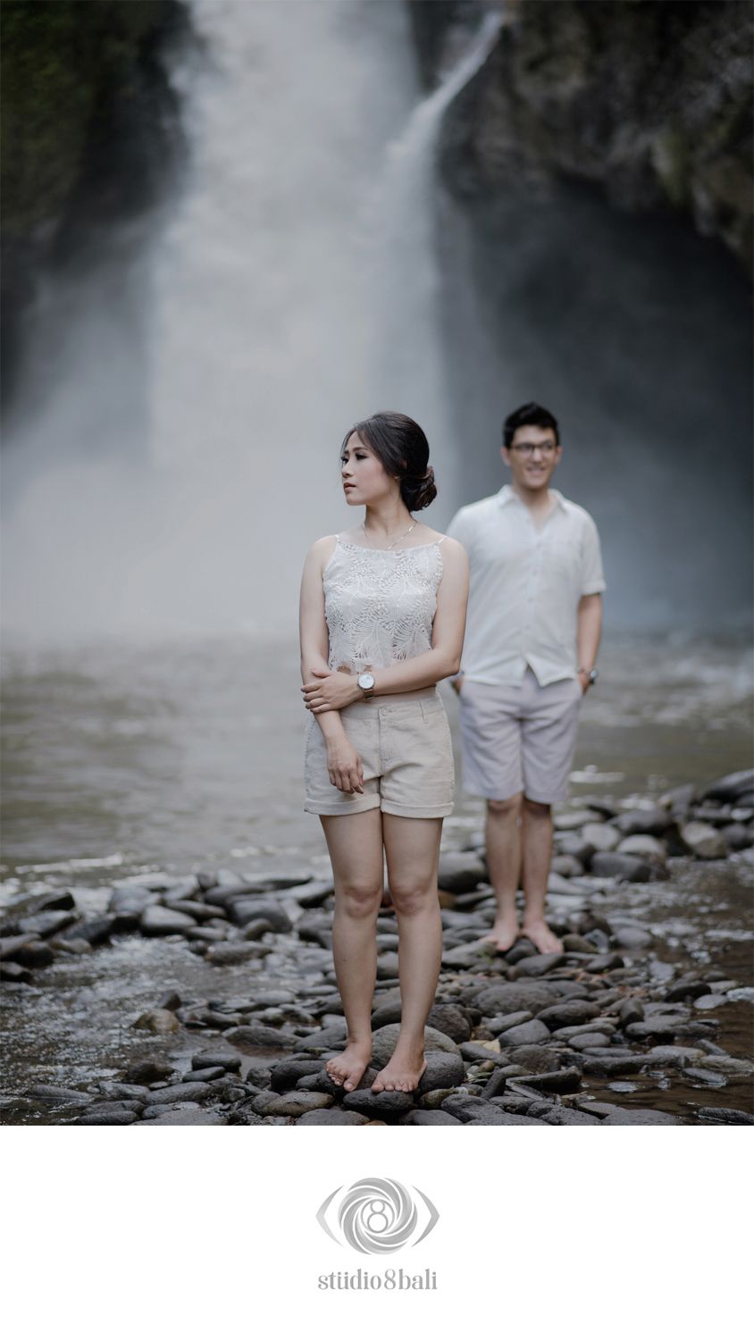 Couple in white outfits posing near a misty waterfall on rocky terrain in Bali, Indonesia.