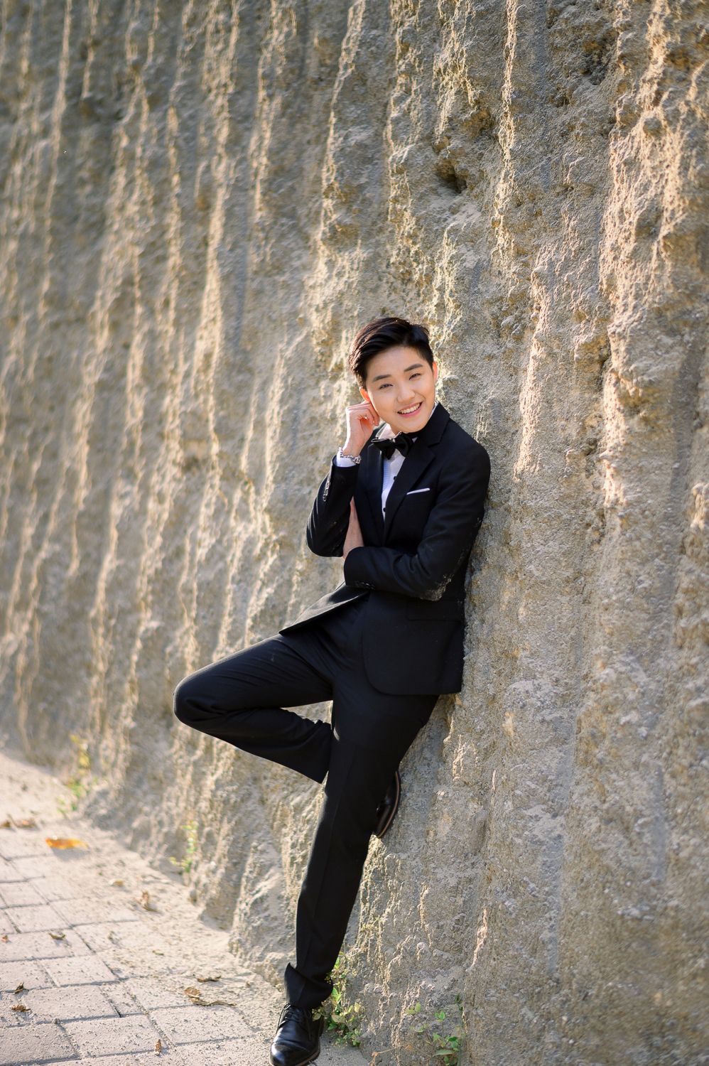 Young Asian man in black tuxedo leaning against stone wall with one leg raised, smiling confidently.