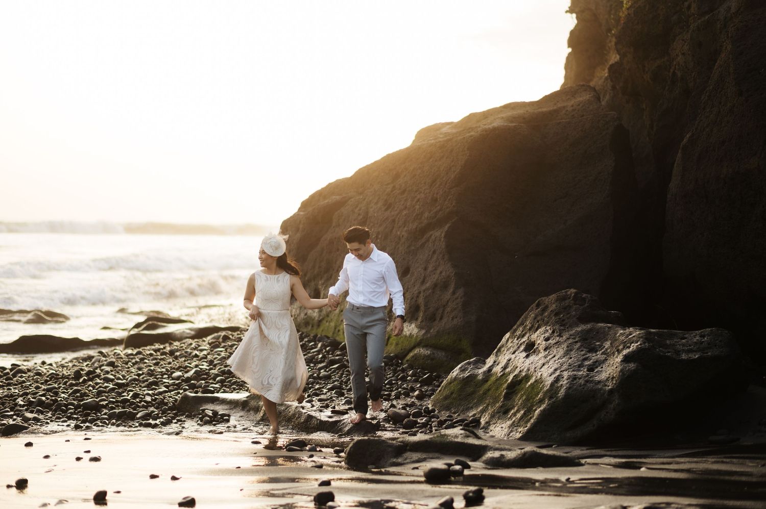Couple holding hands walking on rocky beach at sunset near large cliffs with ocean waves in background.