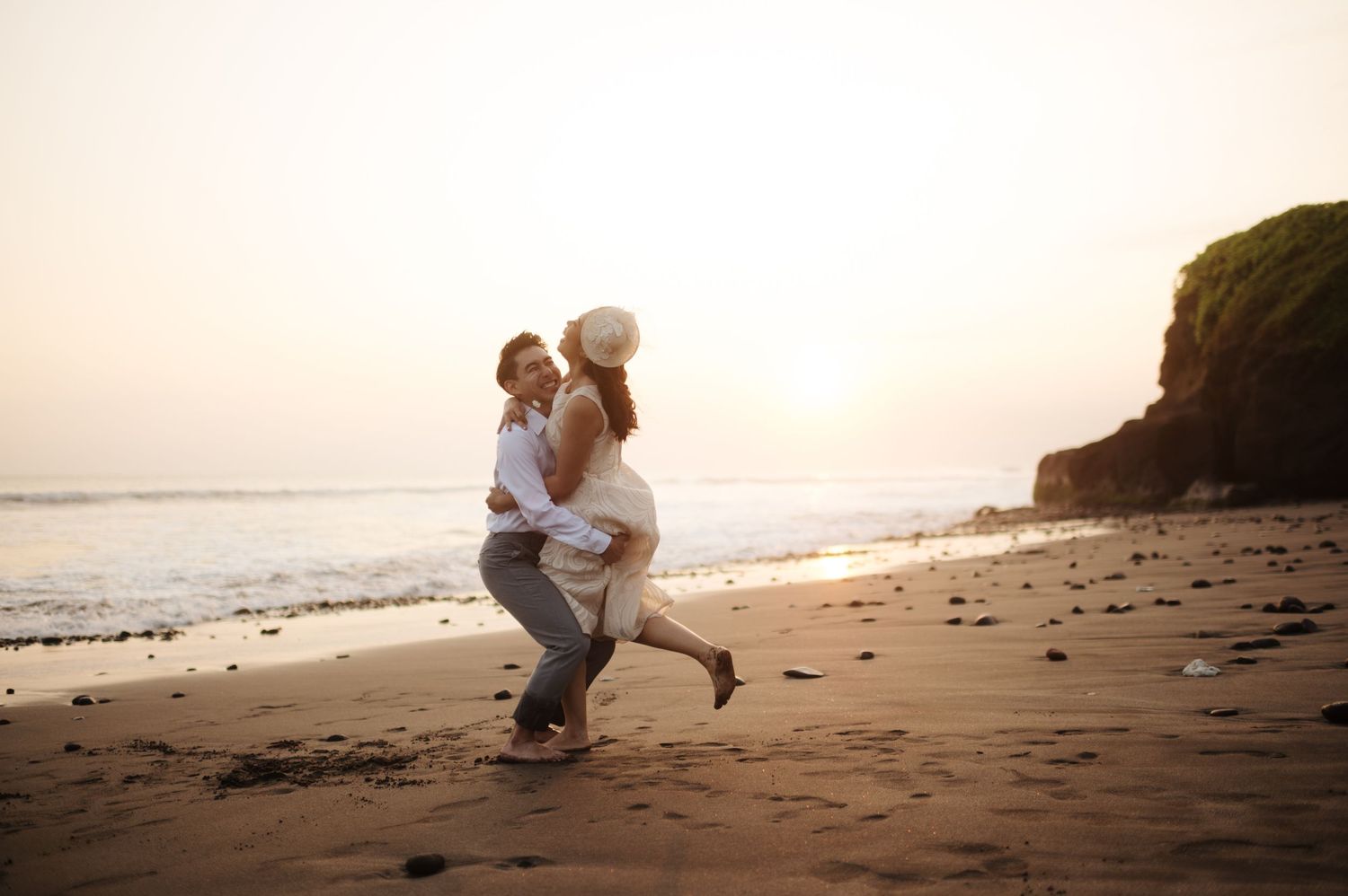 Couple dancing joyfully on a sandy beach at sunset with ocean waves and rocky cliffs in the background.