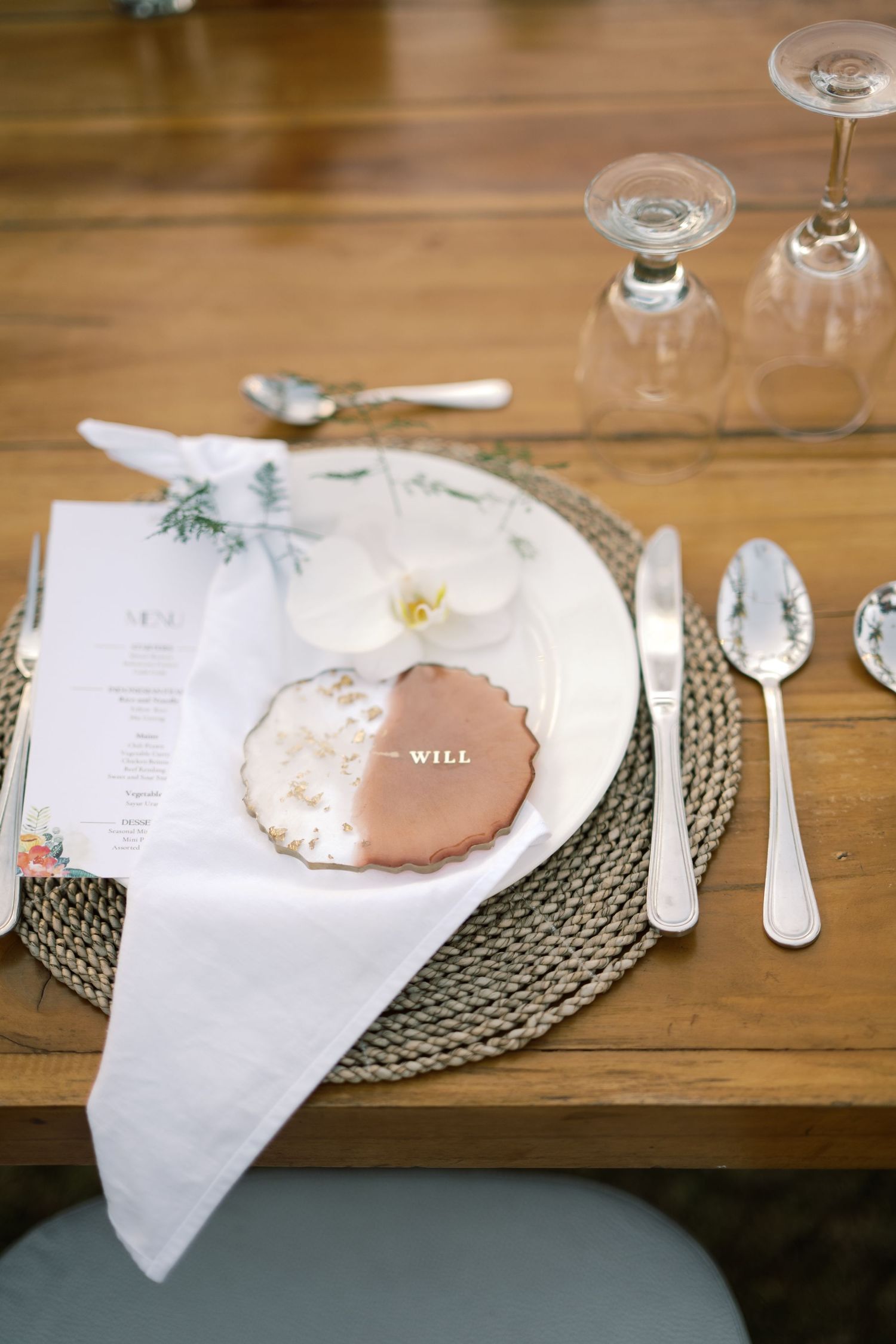 Elegant wedding place setting with floral menu, orchid, personalized cookie name card, silverware, and glasses on woven placemat.