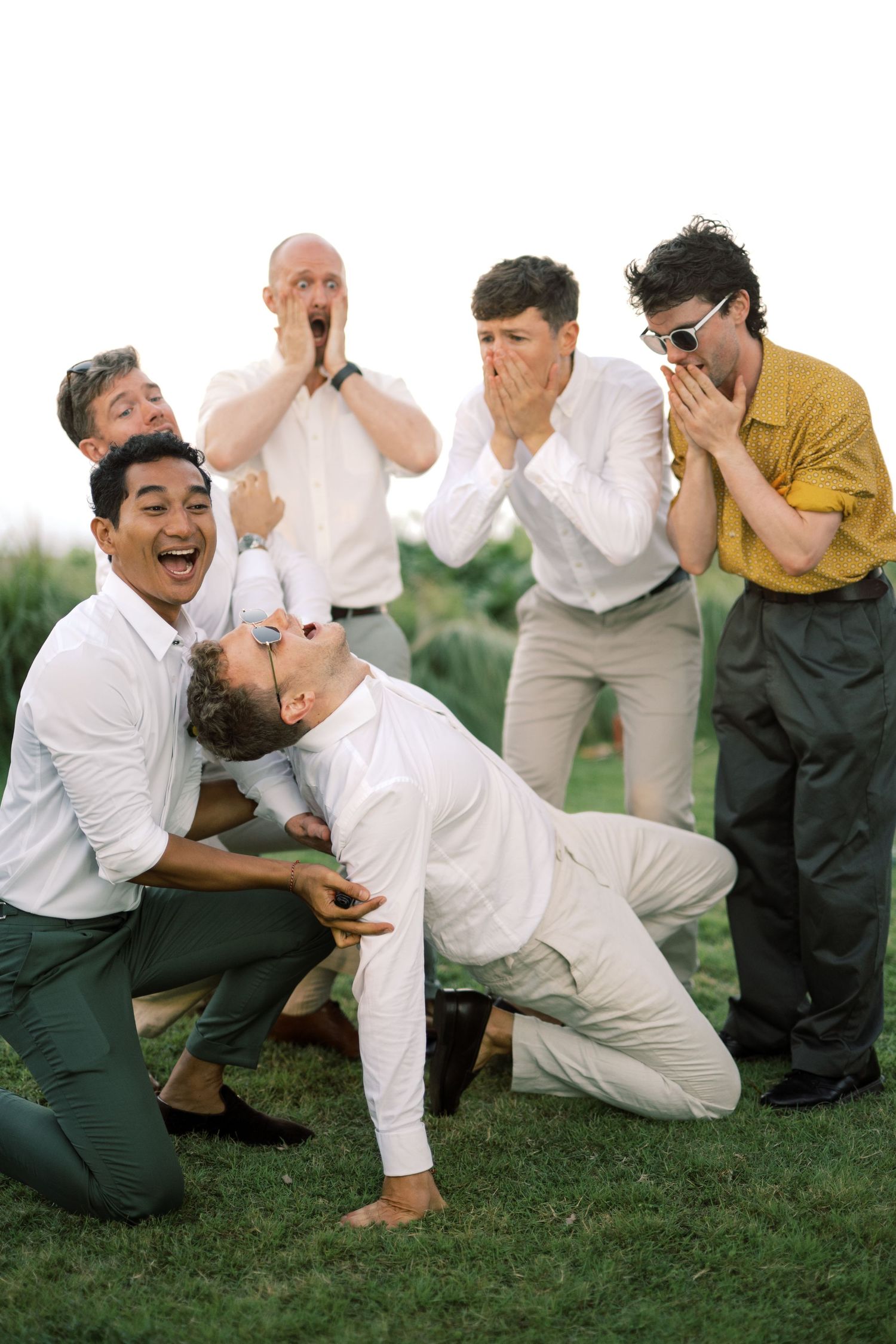 Group of groomsmen reacting with shock and laughter during a fun outdoor wedding party photo on green grass.