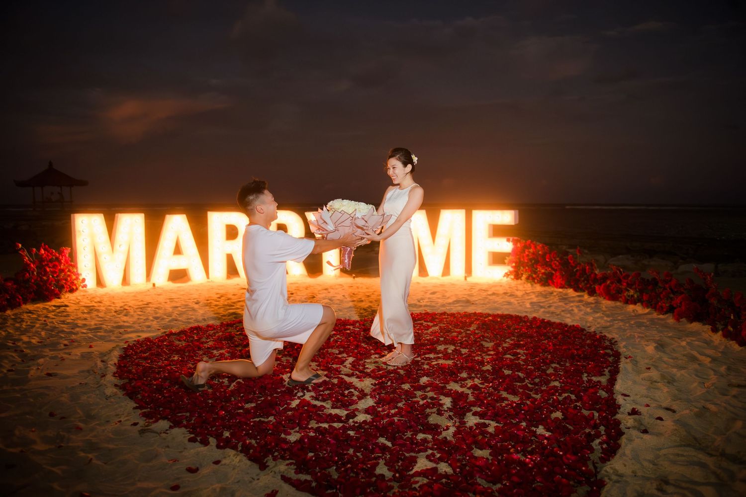 Man proposes on a romantic beach at night with glowing MARRY ME letters and rose petals in a heart shape.