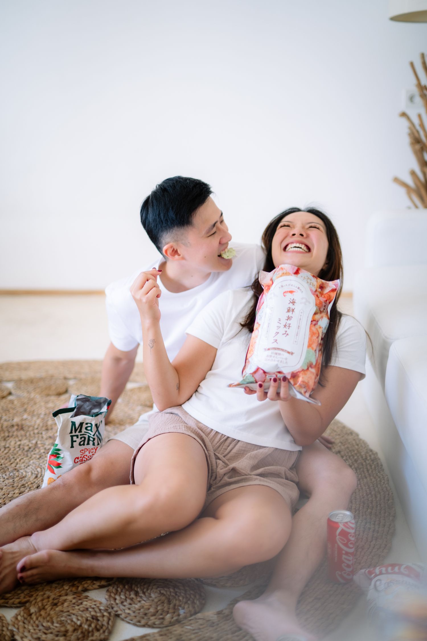 Happy Asian couple sitting on floor in white tees, laughing and snacking from colorful chip bags at home.