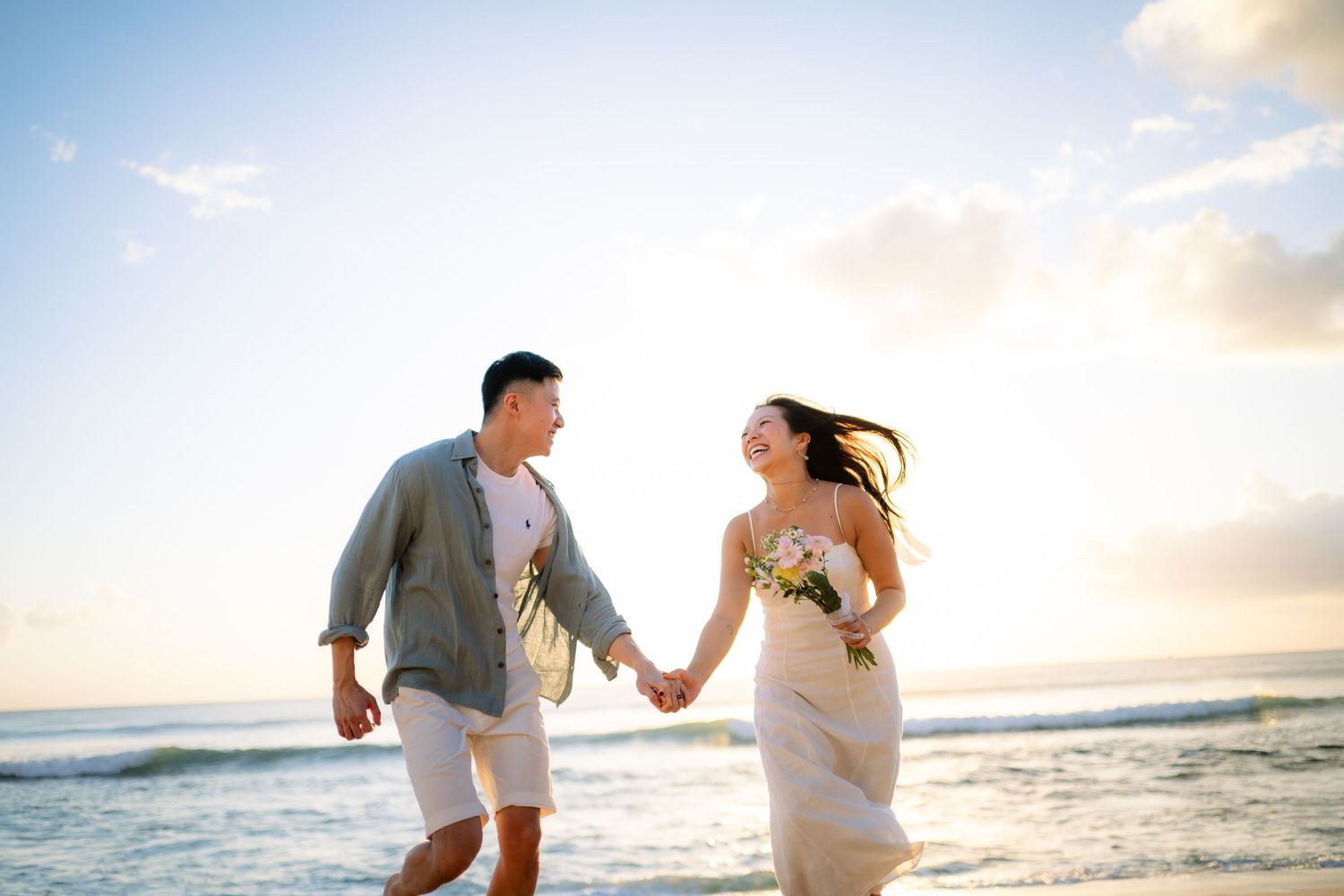 Couple holding hands walking on a sunny beach, woman in white dress with bouquet, man in casual gray shirt.