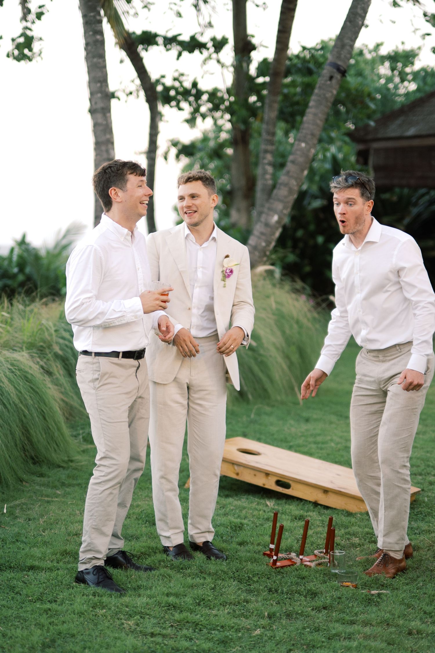 Groom in ivory suit laughing with two groomsmen in white shirts playing lawn games at outdoor wedding.