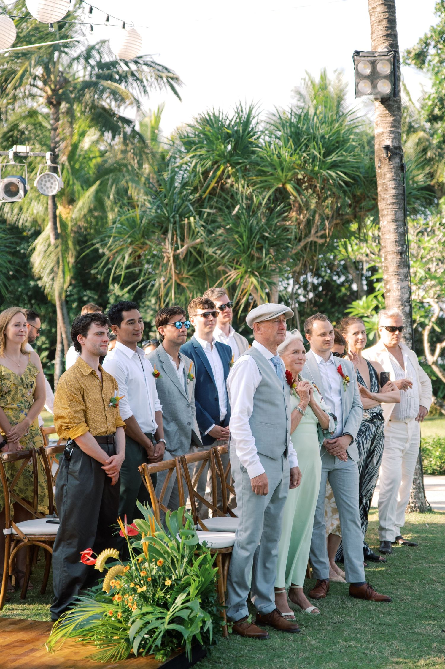 Wedding guests standing outdoors at tropical ceremony surrounded by palm trees and colorful floral arrangements.