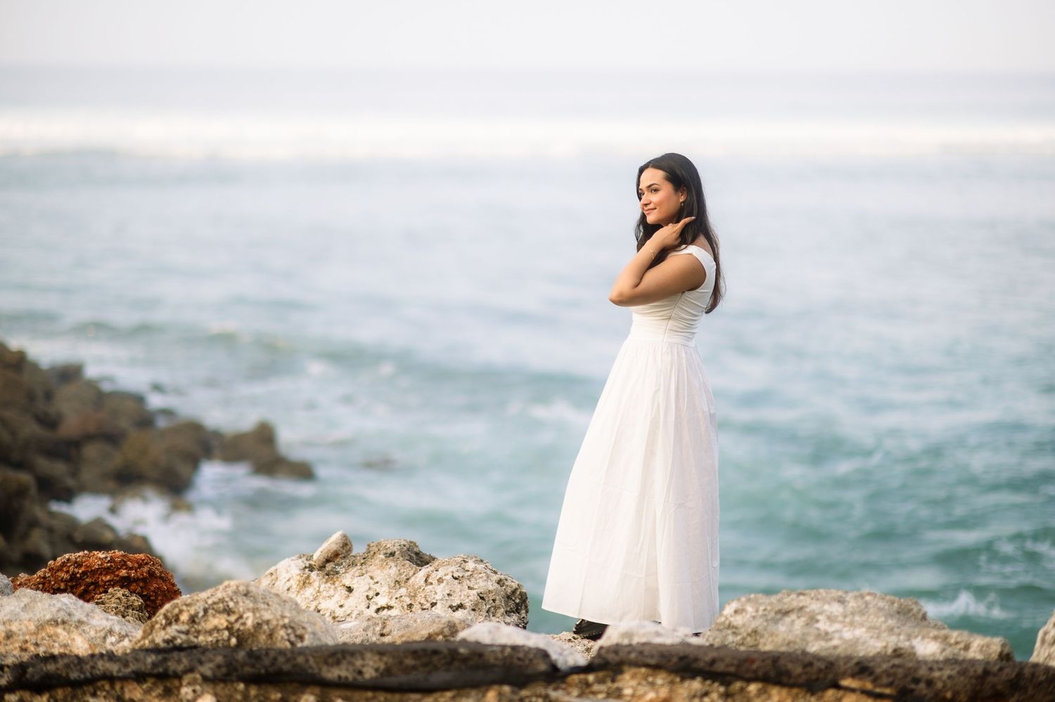 A woman in a white dress stands on rocky coastal cliffs with ocean waves in the background.