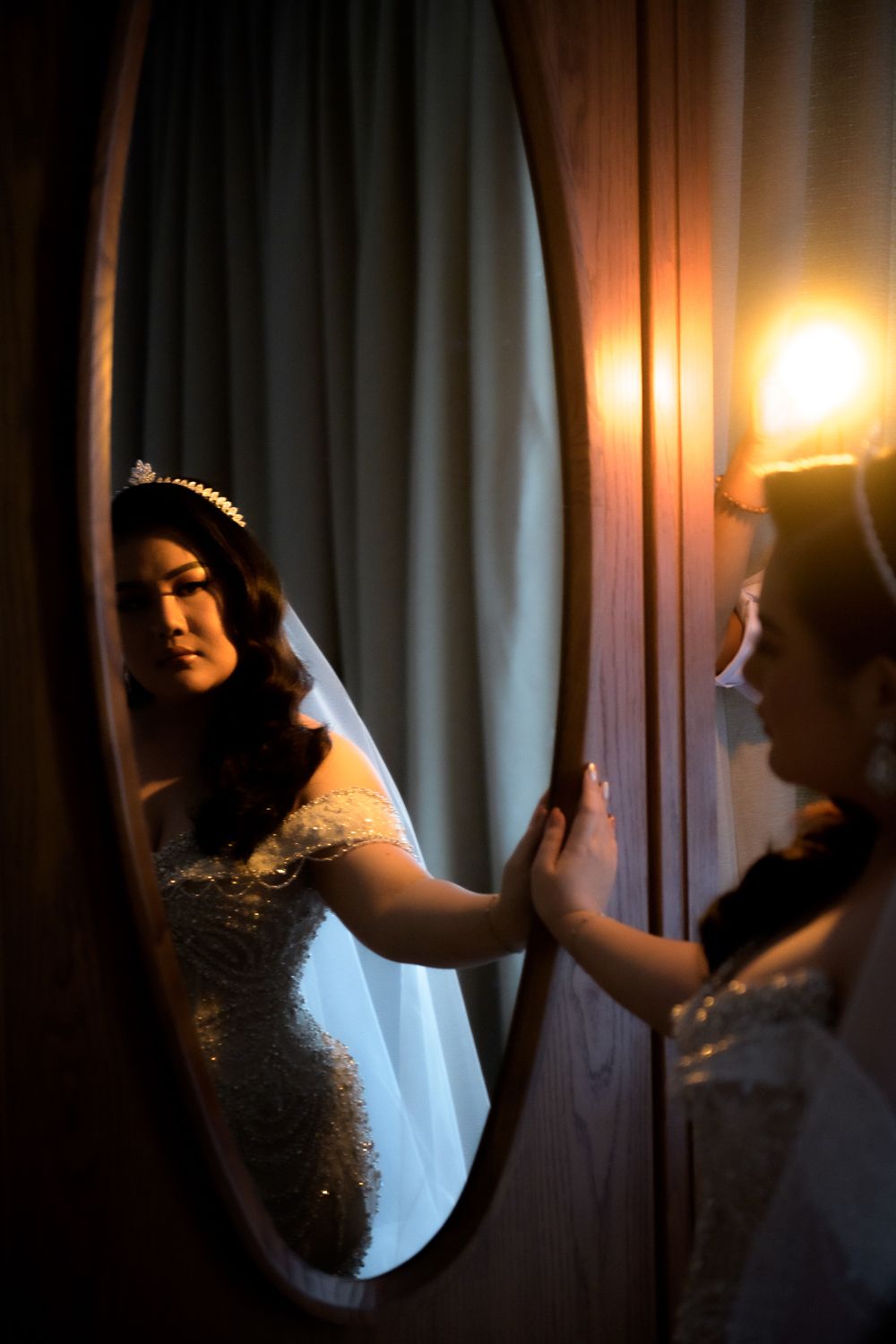 Bride in a beaded wedding gown and tiara gazes at her reflection in an oval mirror, illuminated by warm vanity lighting.