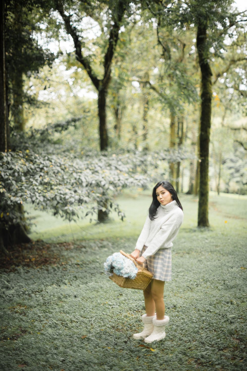 Young woman in white sweater and plaid skirt holds a wicker basket with flowers in a lush green forest.