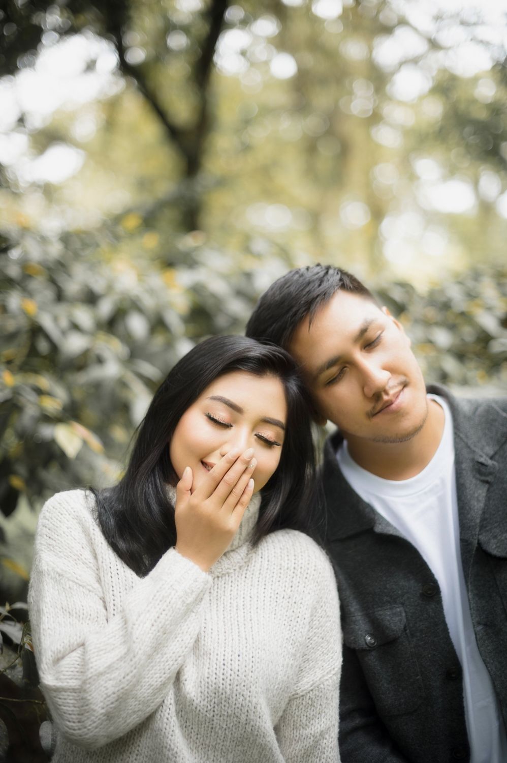 Young Asian couple with eyes closed sharing a tender moment outdoors, woman in white sweater, man in dark jacket.