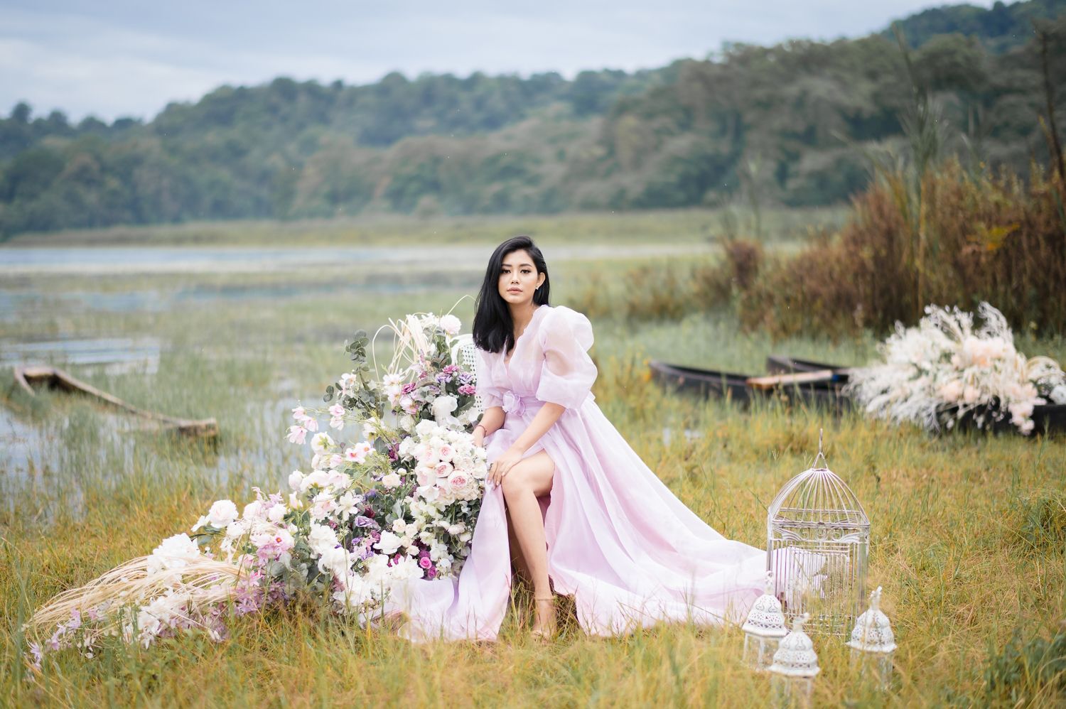 Woman in pink gown poses with flowers by a serene lake surrounded by lush greenery.