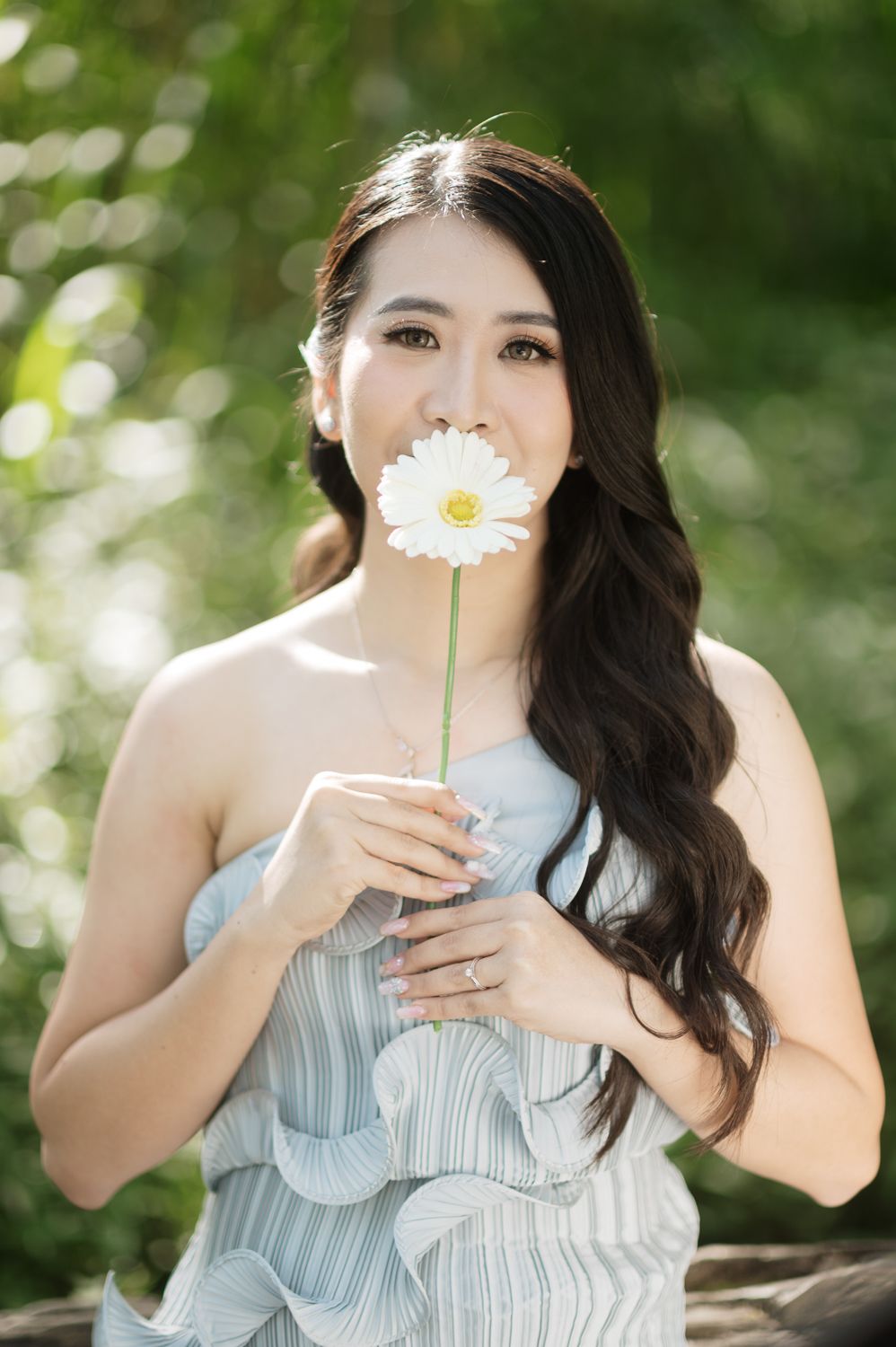 A young Asian woman in a blue dress holds a white daisy flower near her face outdoors.