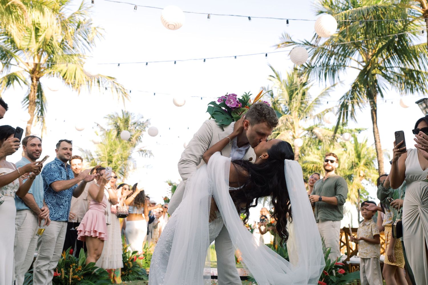 Groom dips bride in a romantic kiss as wedding guests cheer at a tropical outdoor reception.