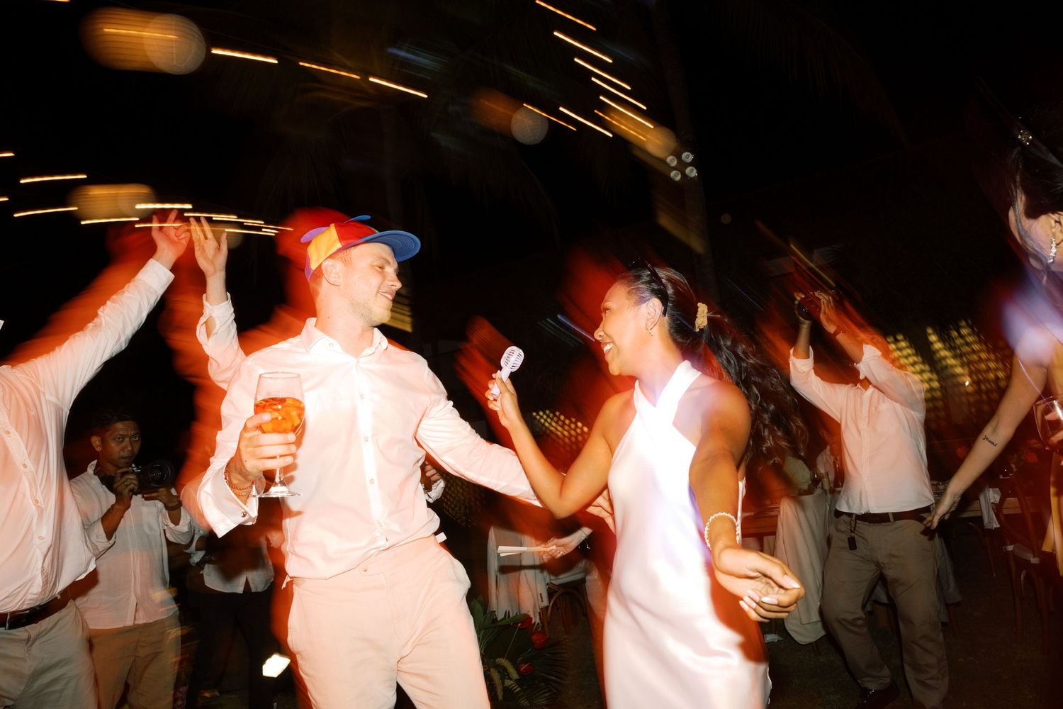 Bride and groom dance energetically with guests under colorful lights at lively outdoor tropical wedding reception.
