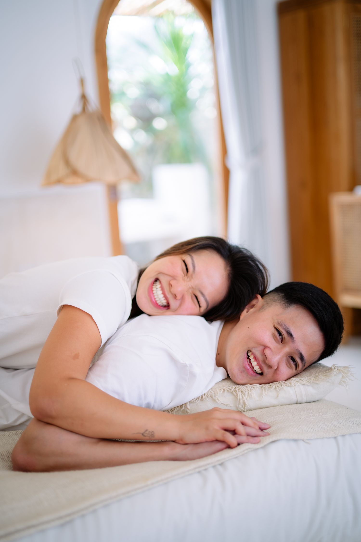 Happy couple lying on a bed, smiling and laughing together in a bright, cozy bedroom with white bedding.