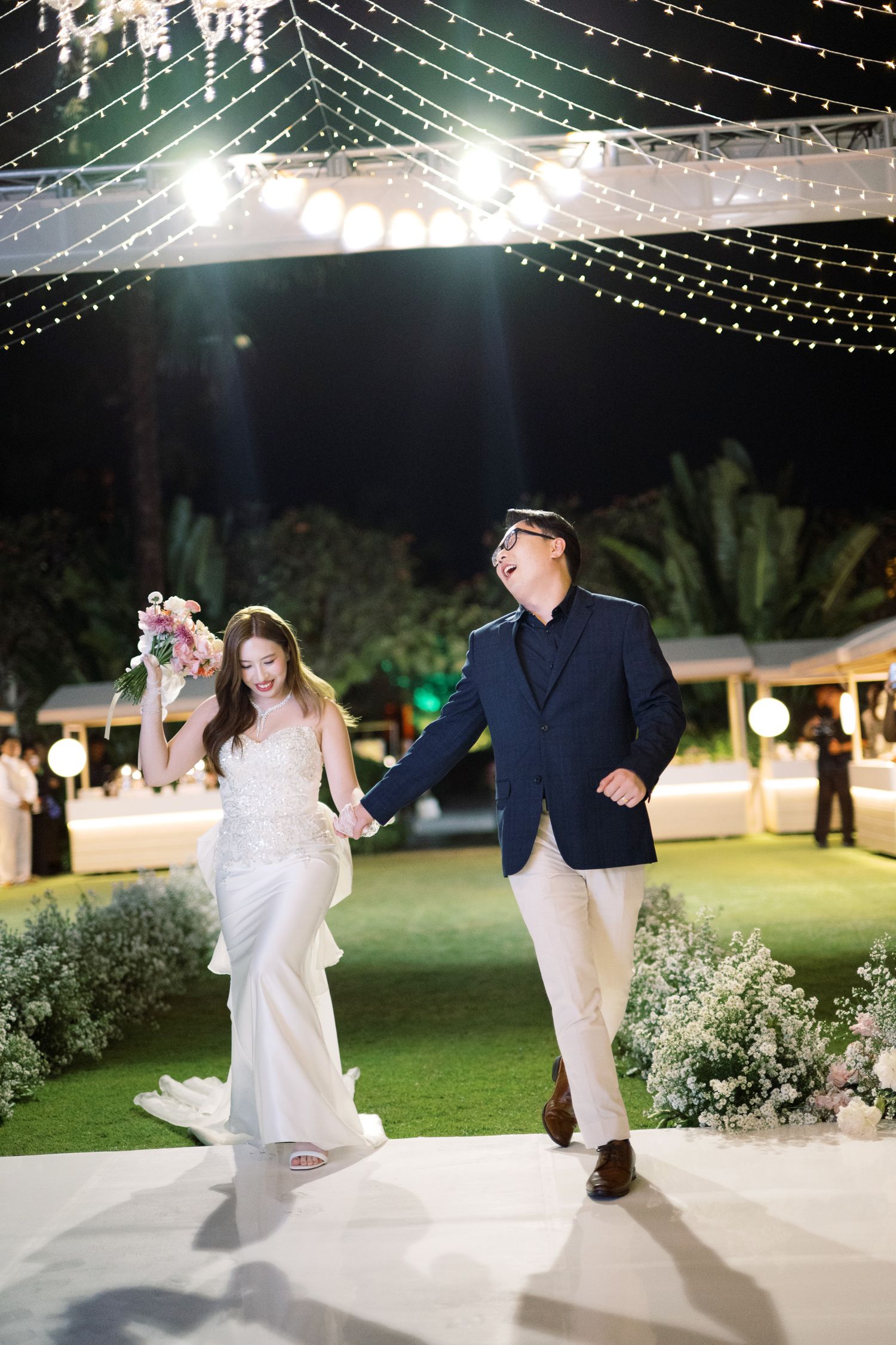 Bride in white gown and groom in navy suit dancing joyfully at an outdoor evening wedding reception with string lights.