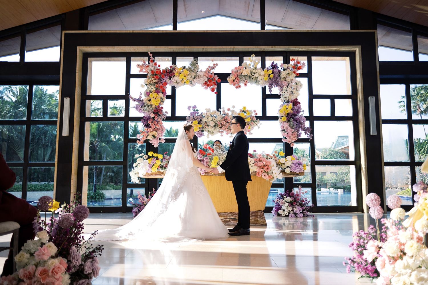Bride and groom exchange vows at floral arch altar in elegant sunlit wedding chapel with colorful flower arrangements.