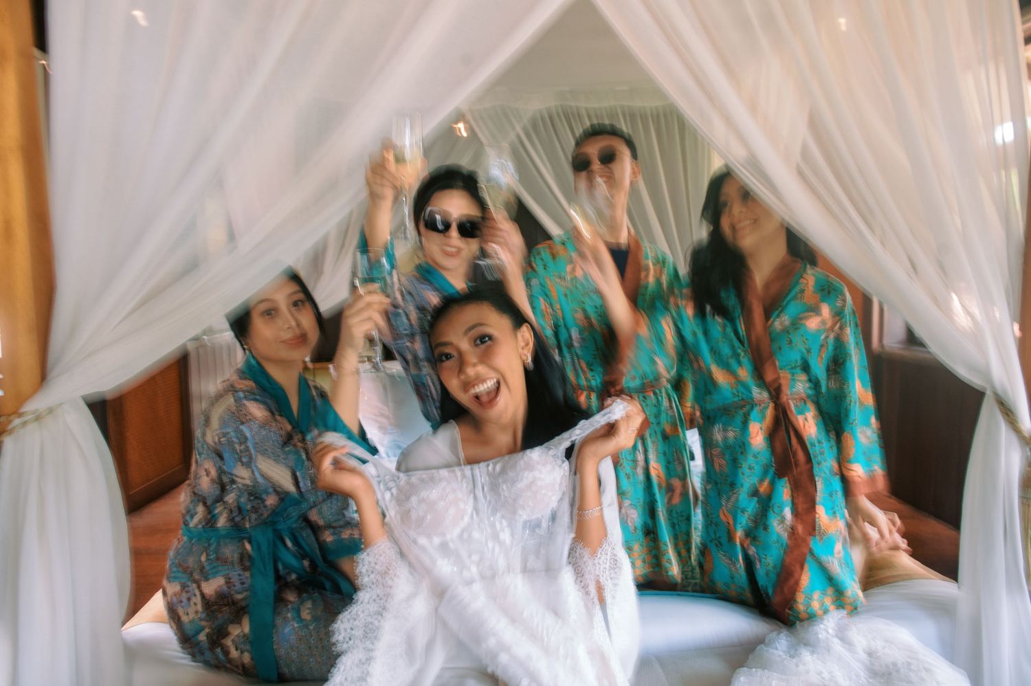 Bride in white robe surrounded by bridesmaids in teal floral robes celebrating on a canopy bed.