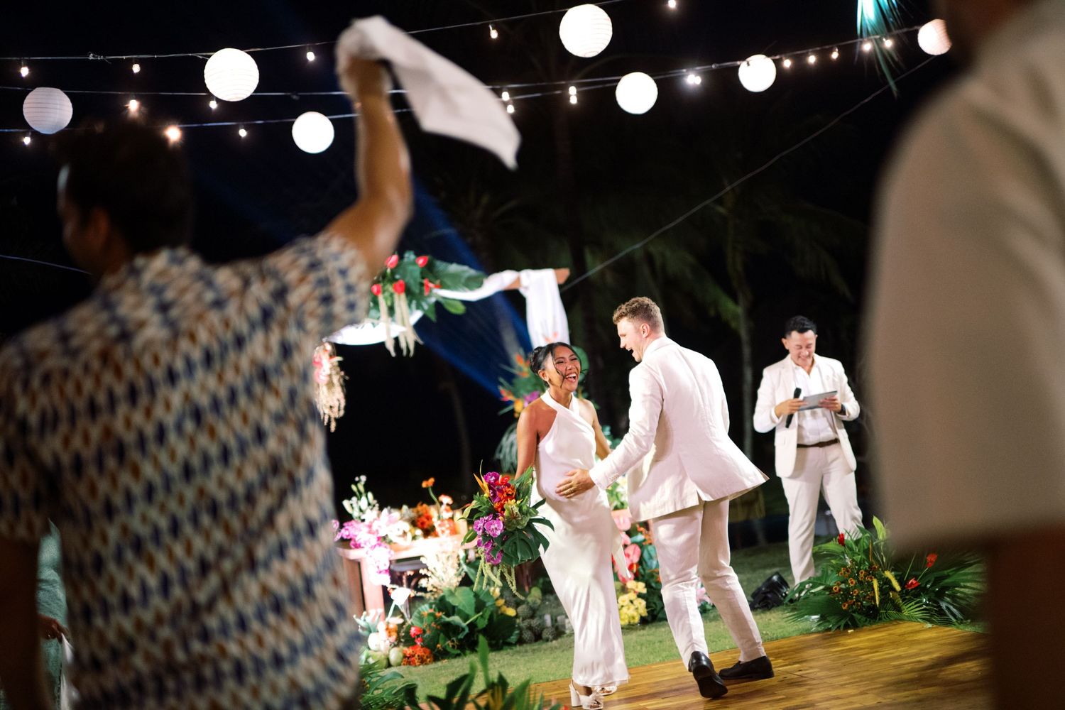 Outdoor tropical wedding ceremony at night with string lights, bride and groom exchanging vows on decorated stage.