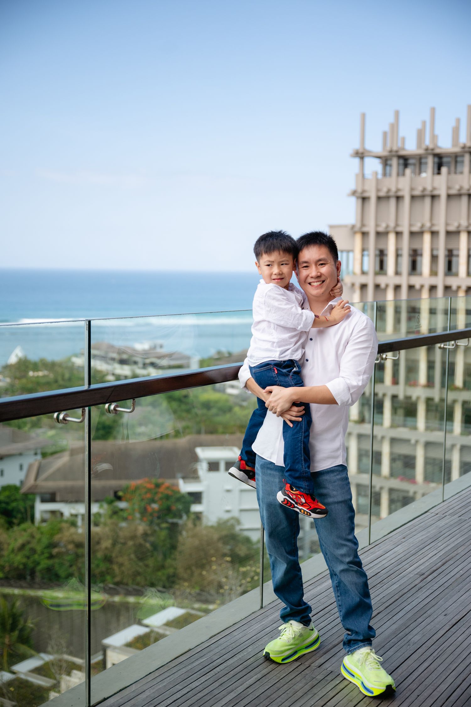 Asian father holding young son on a balcony with ocean and city views on a sunny day.
