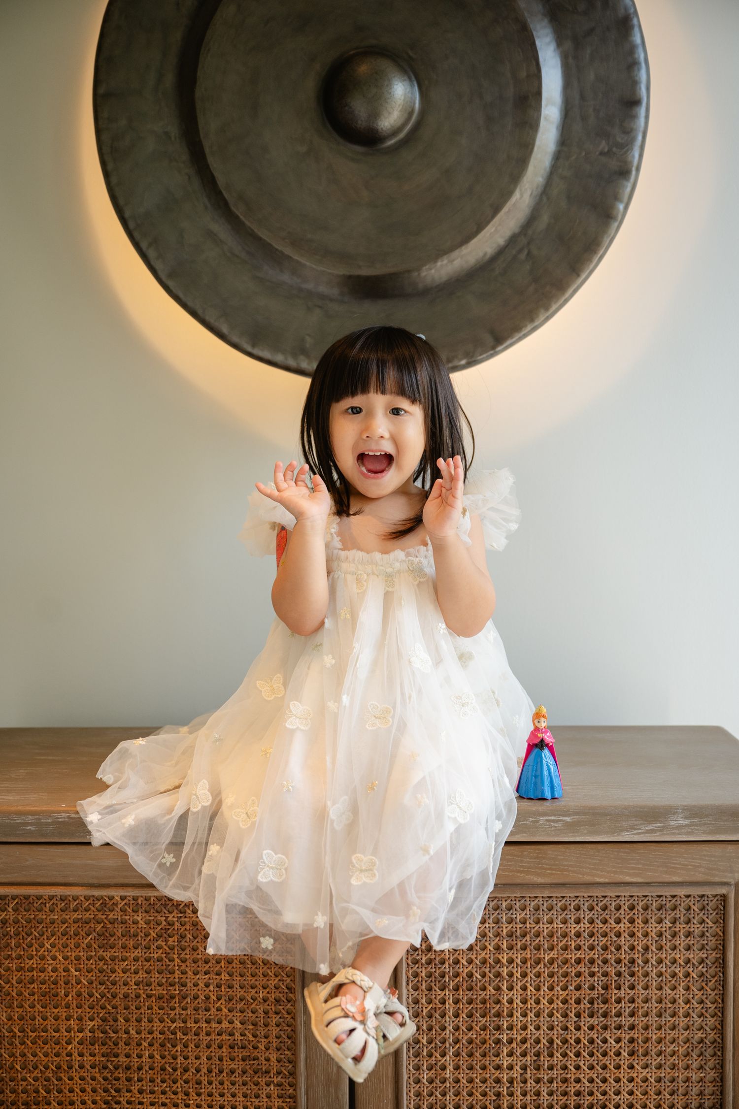 Excited young girl in white tulle dress sitting on rattan cabinet, hands raised, with decorative gong backdrop.