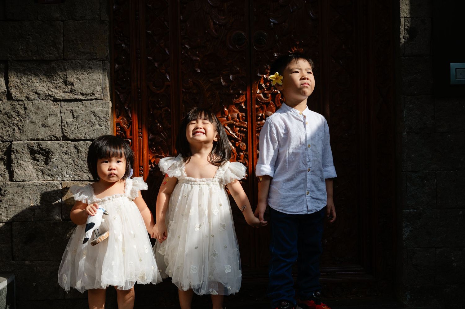 Three Asian children in white dresses and blue shirt hold hands smiling outdoors near ornate wooden doors.