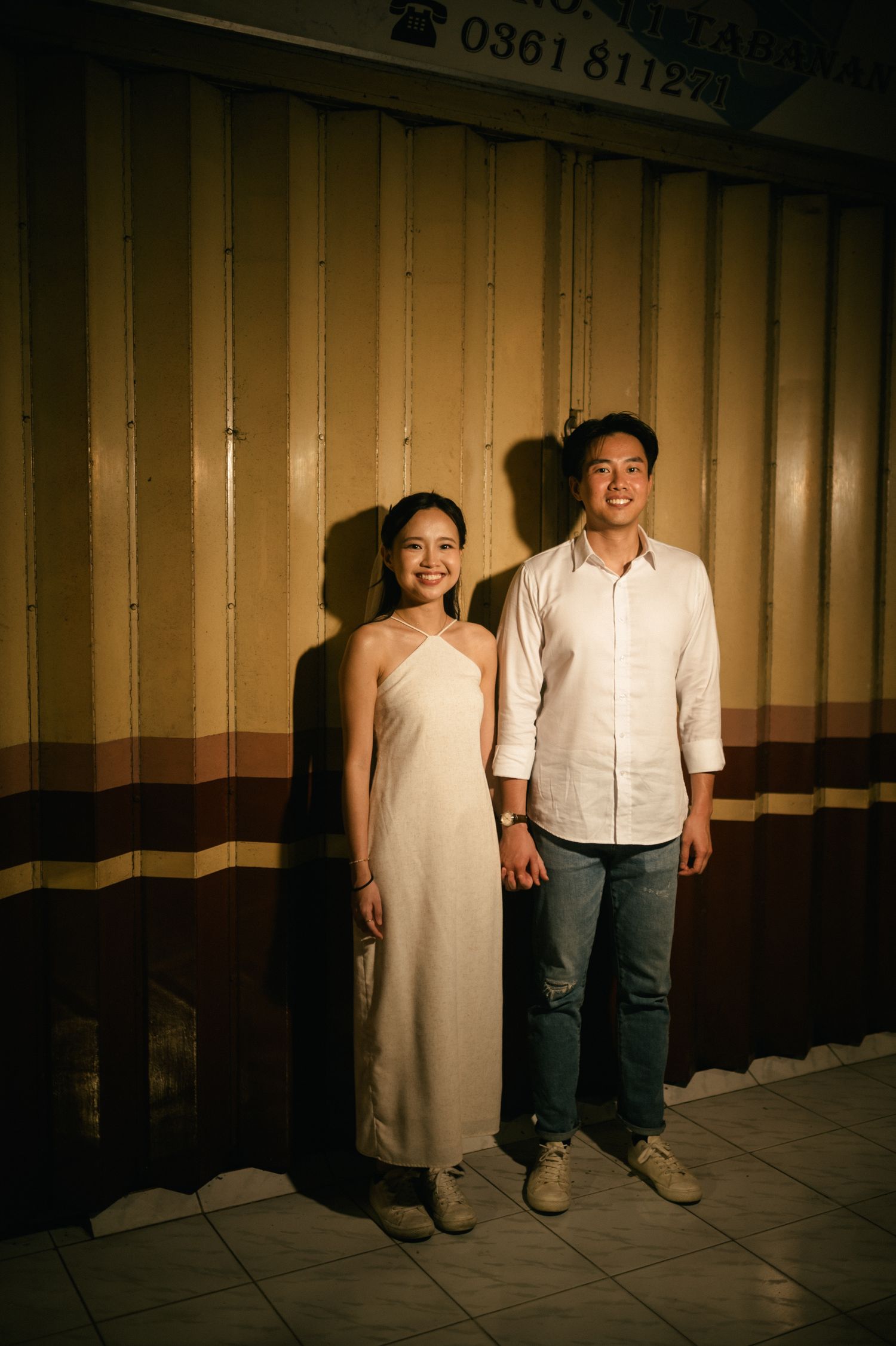 A smiling couple in elegant attire poses together against a striped wall at night.