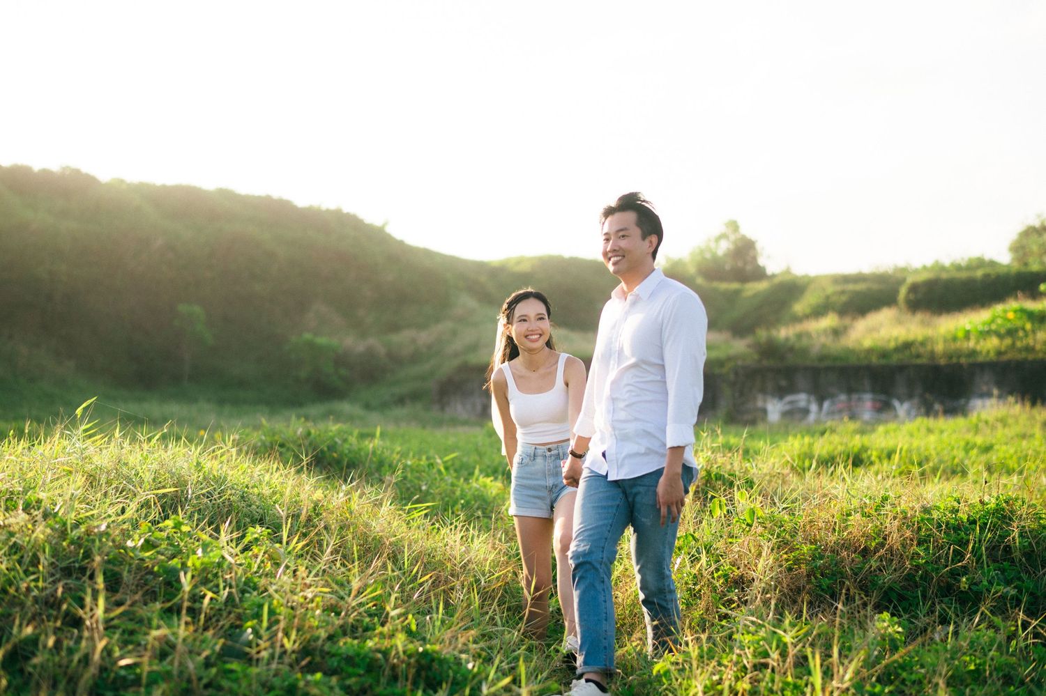 Happy couple walking hand in hand through lush green field during golden hour sunset outdoors.