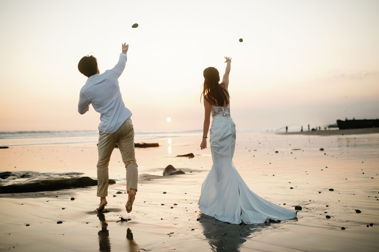 Bride and groom tossing rocks into the ocean at sunset during a beach wedding photoshoot.