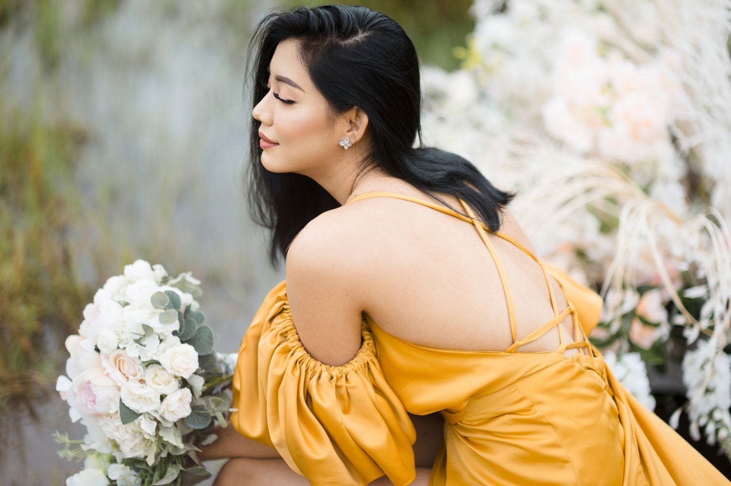 Woman in yellow off-shoulder satin dress holding white bouquet in elegant outdoor setting.