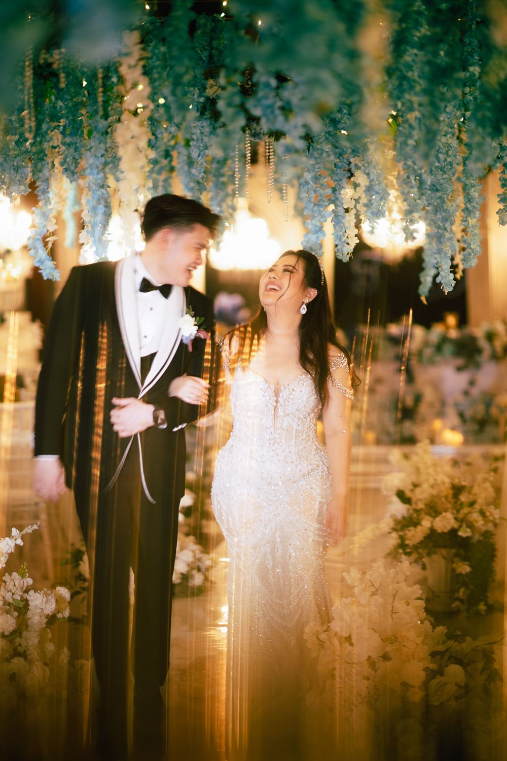 Bride in beaded gown laughs joyfully with groom in tuxedo under hanging floral decor at elegant wedding reception.
