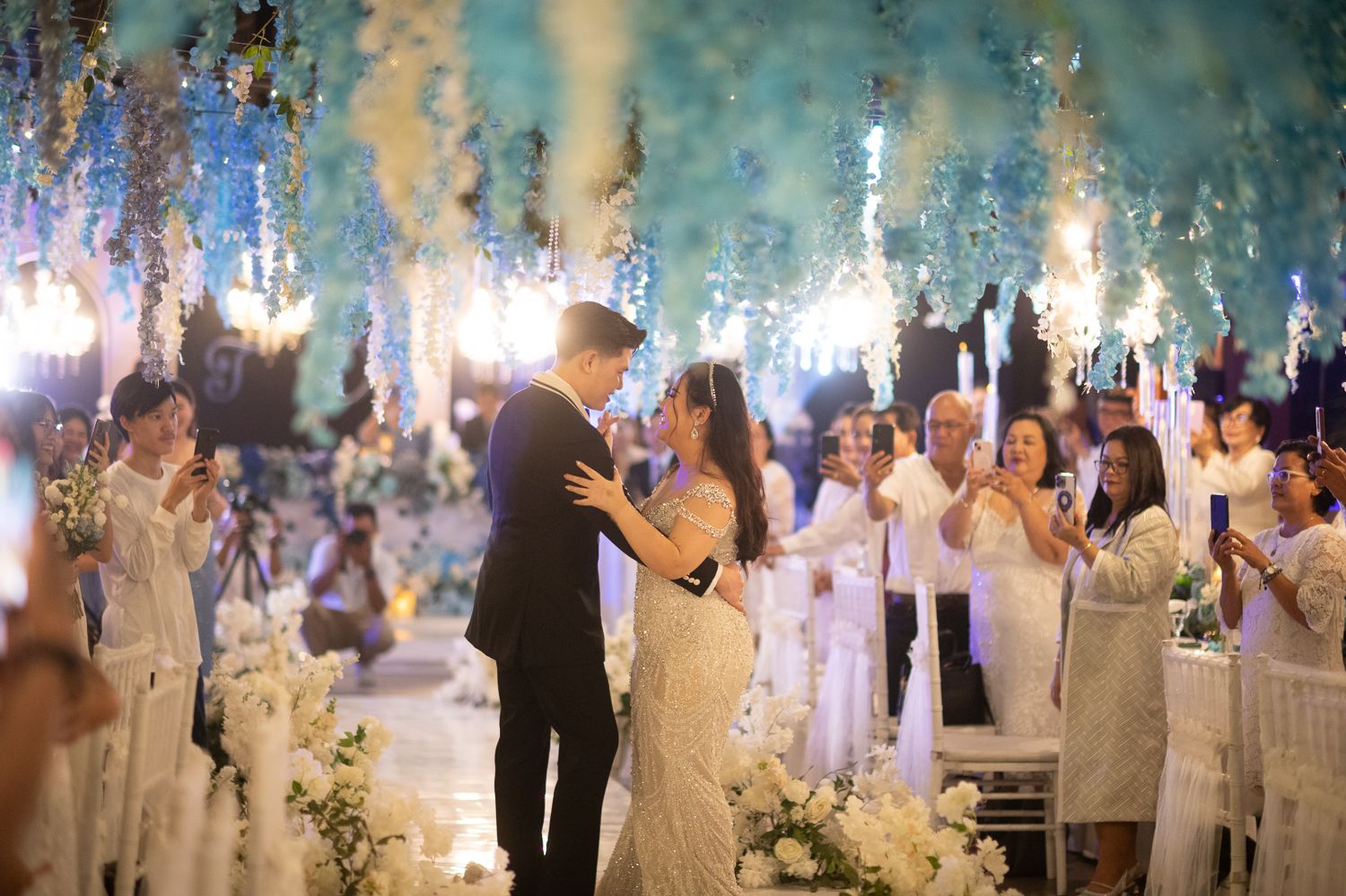 Couple shares first dance at elegant wedding reception with white floral decor, sparkling lights, and guests in white attire.
