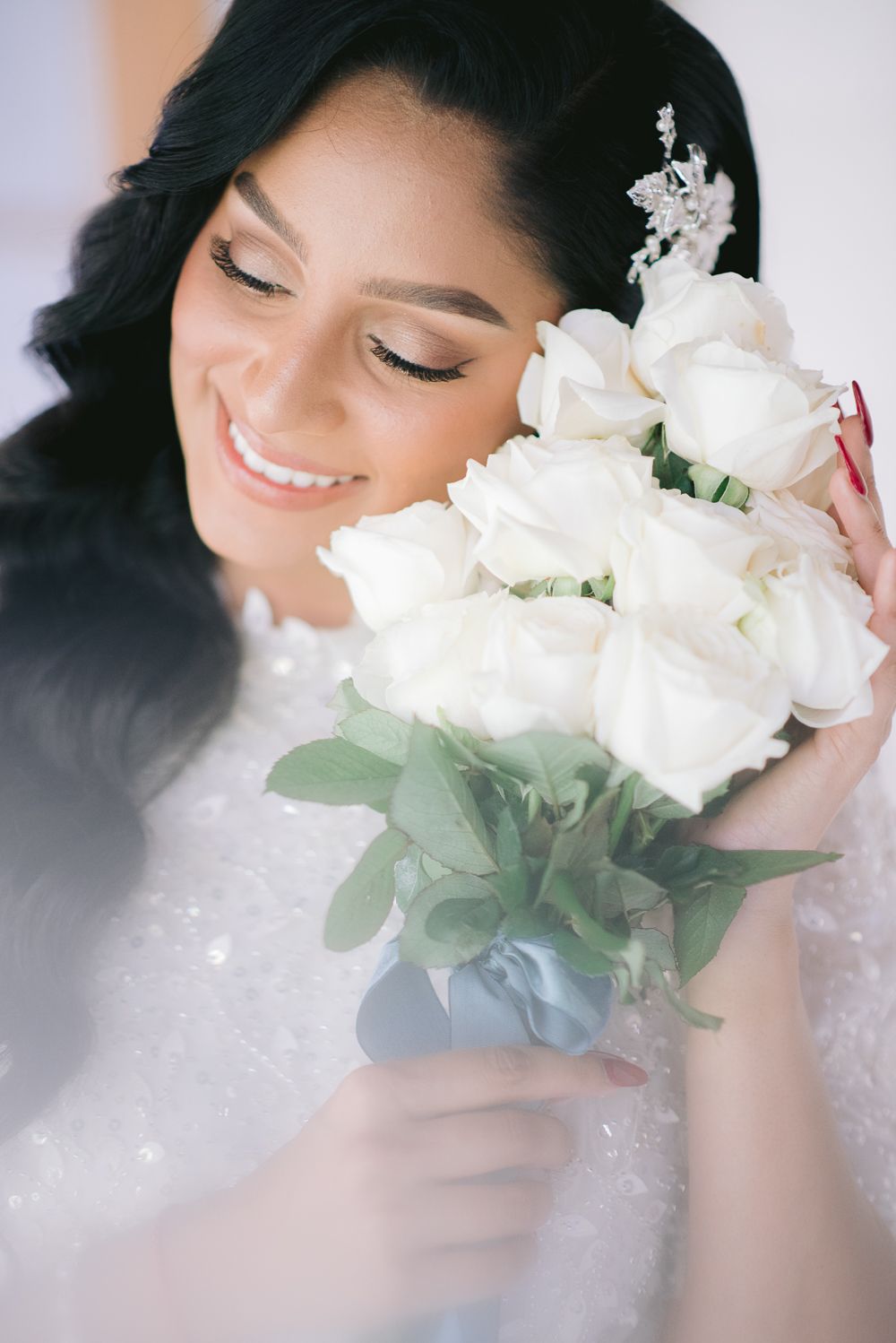 Smiling bride with dark hair holds white rose bouquet close to her face, wearing a sparkly wedding gown.