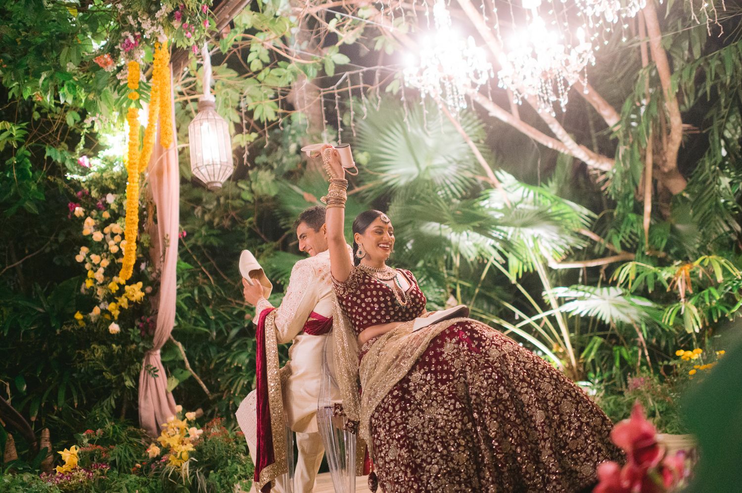 Bride in burgundy lehenga celebrates at outdoor Indian wedding ceremony surrounded by lush greenery and fairy lights.