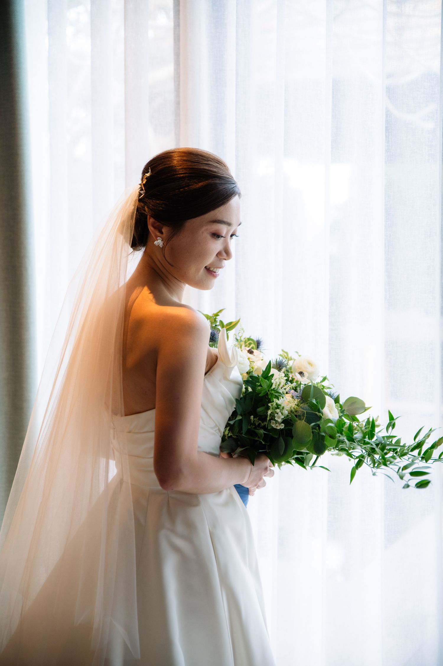 Bride in a strapless white gown and long veil holds a green and white bouquet while smiling softly beside a sunlit window.