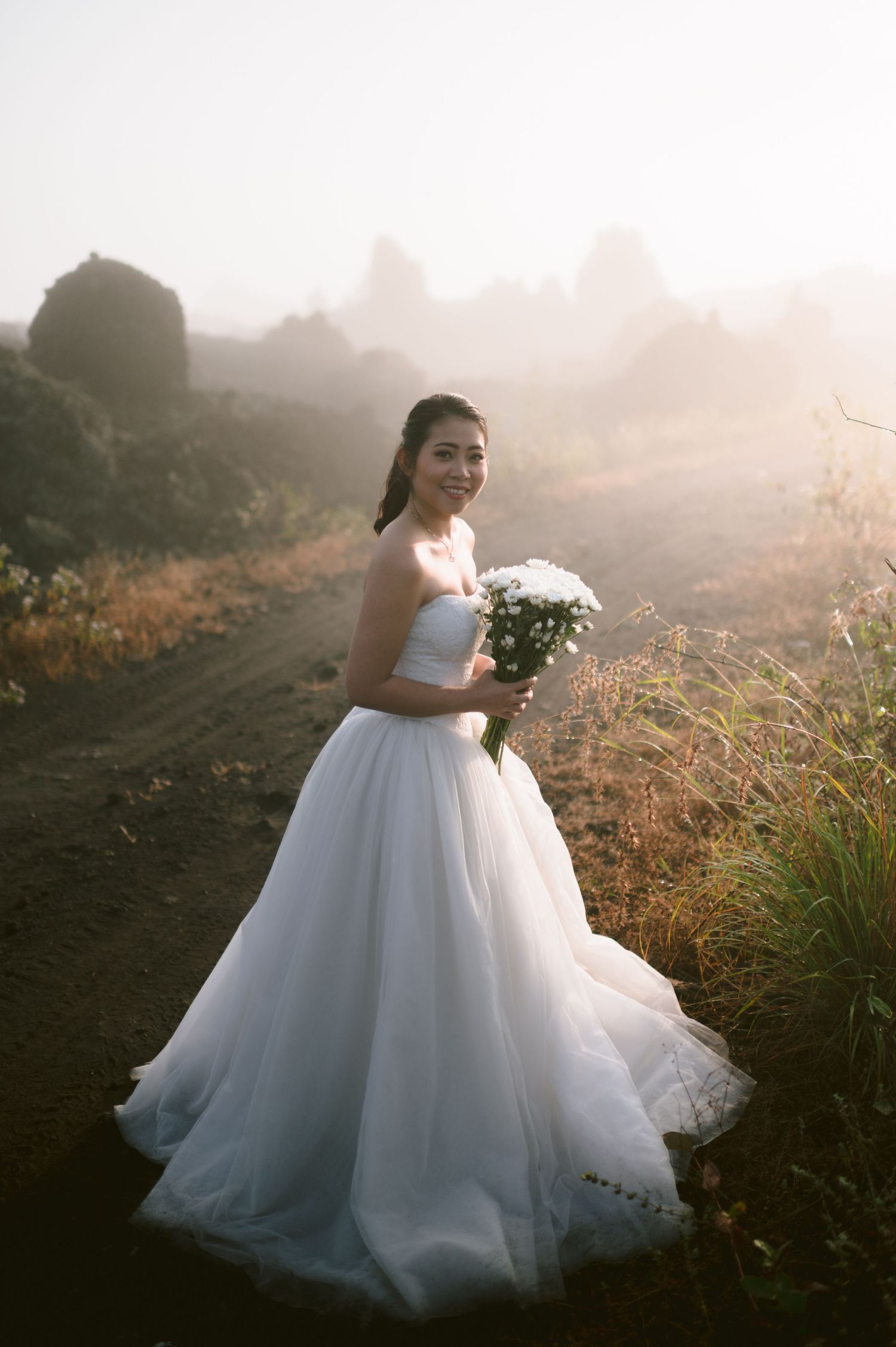 Smiling bride in white ball gown holding bouquet in misty outdoor setting.