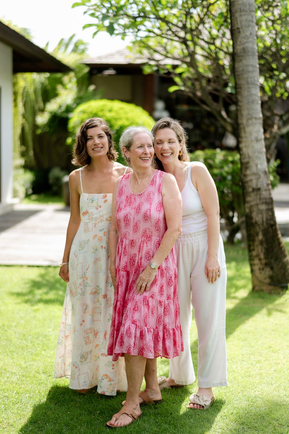 Three women smiling and posing together outdoors on green grass, wearing summer dresses in a tropical garden setting.