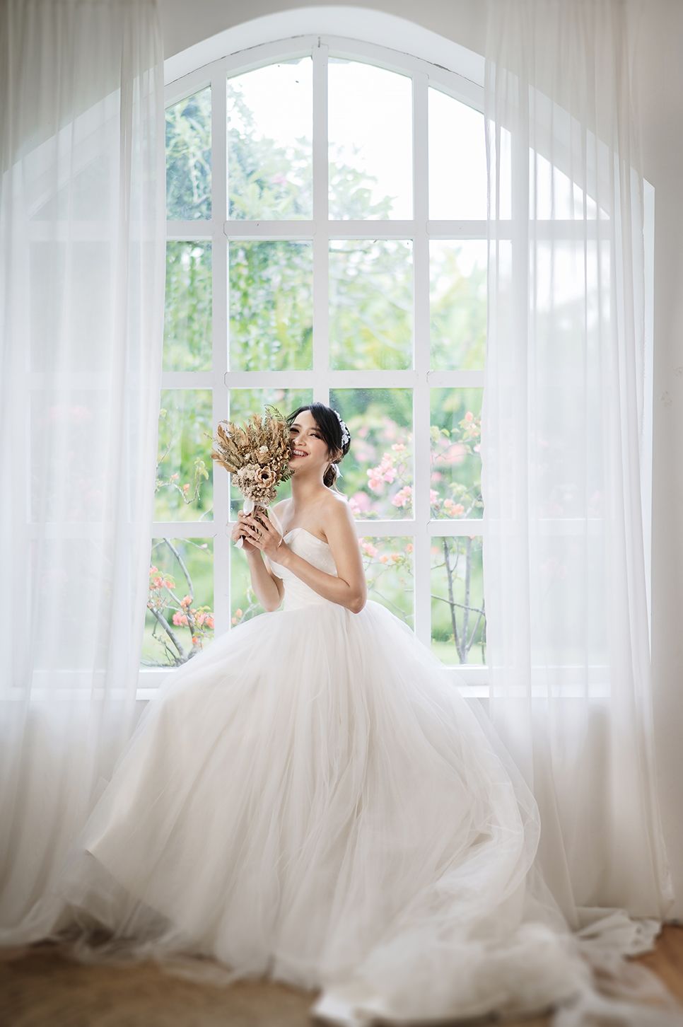 Smiling bride in a white strapless ballgown holds a dried bouquet by an arched window with sheer curtains.