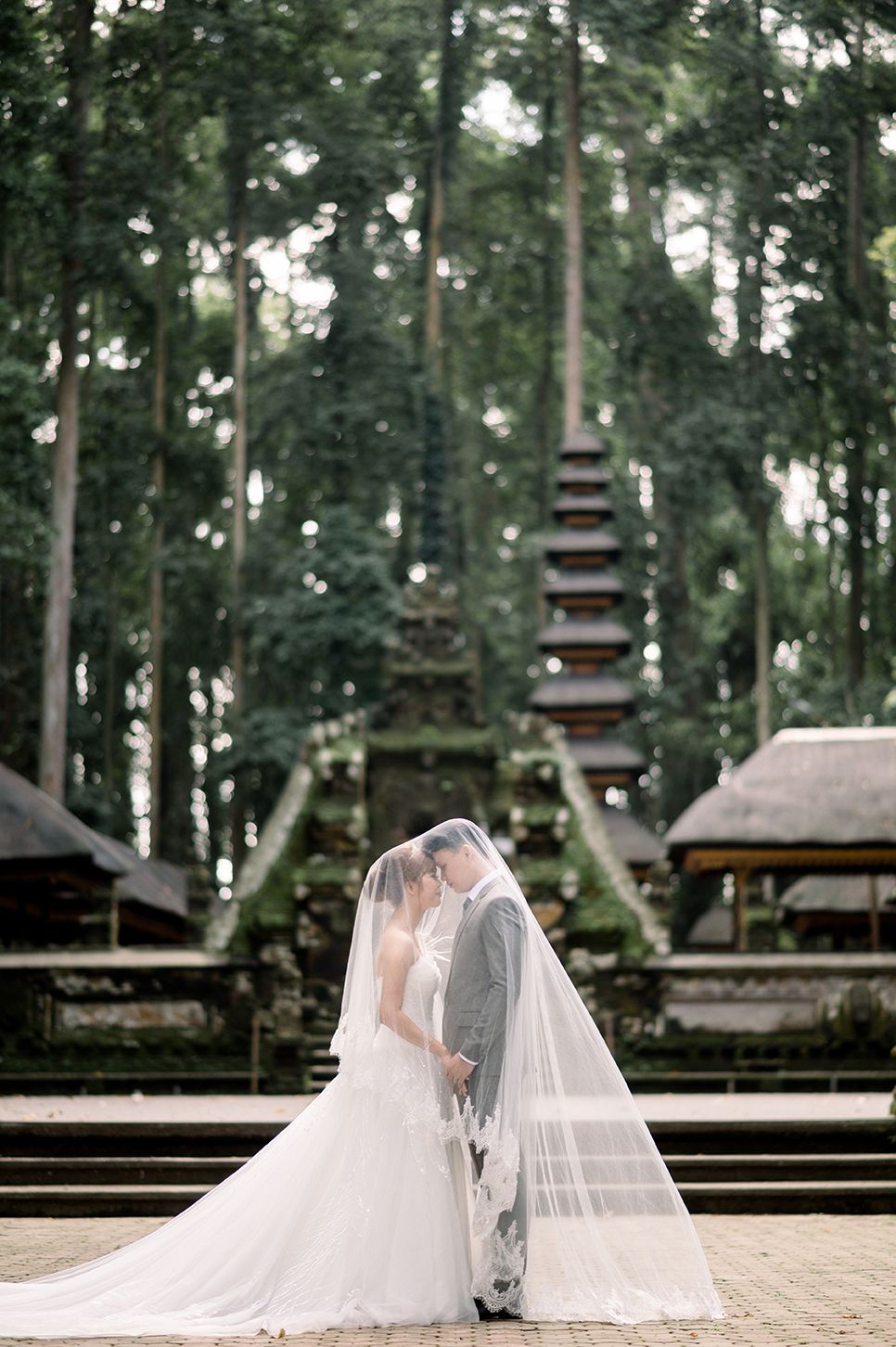Bride and groom share a kiss under a veil before a Balinese temple surrounded by tall trees.