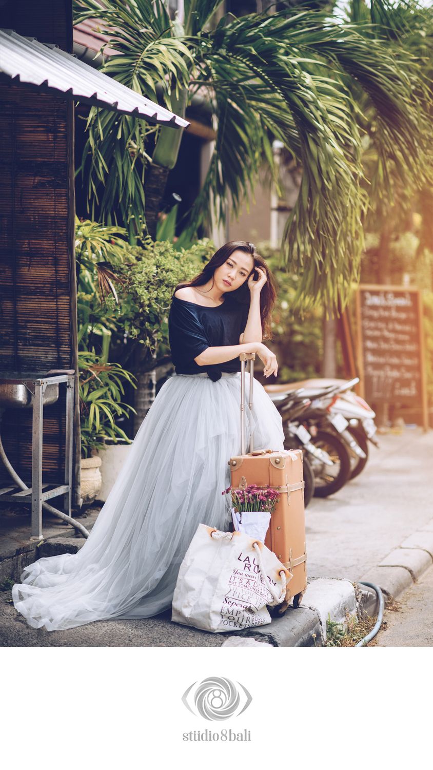 Young woman in black top and gray tulle skirt poses outdoors with vintage suitcase and tote bag near tropical plants.