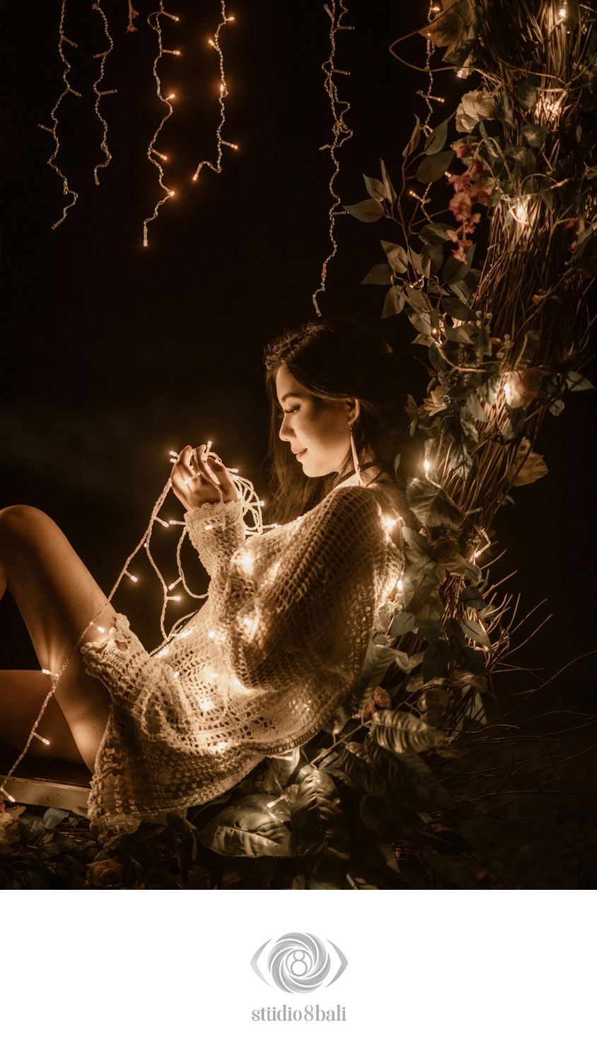 Young woman in crochet sweater wrapped in fairy lights sits against floral backdrop in moody dark setting.