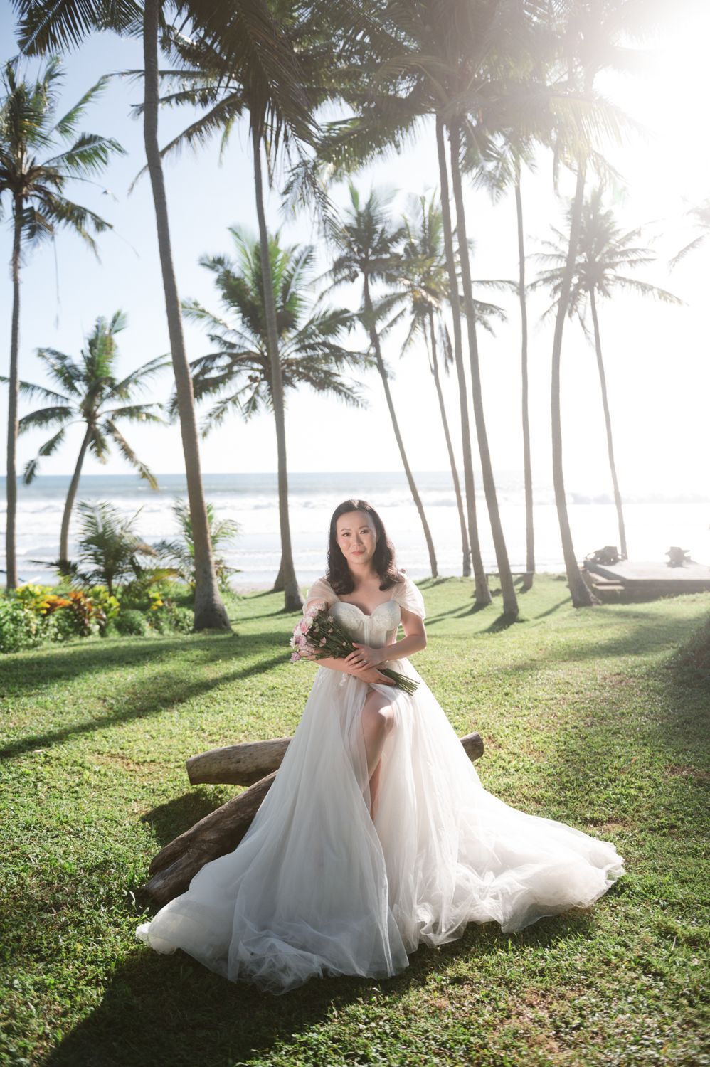 Bride in elegant white tulle gown holding bouquet under tropical palm trees near the beach.