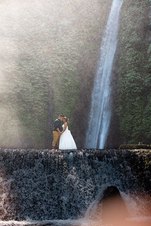 Couple kissing in wedding attire on a stone ledge with a stunning waterfall and lush greenery backdrop.
