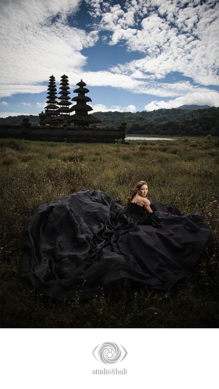 Woman in black gown poses before Balinese temple pagodas in lush green field under dramatic cloudy sky.