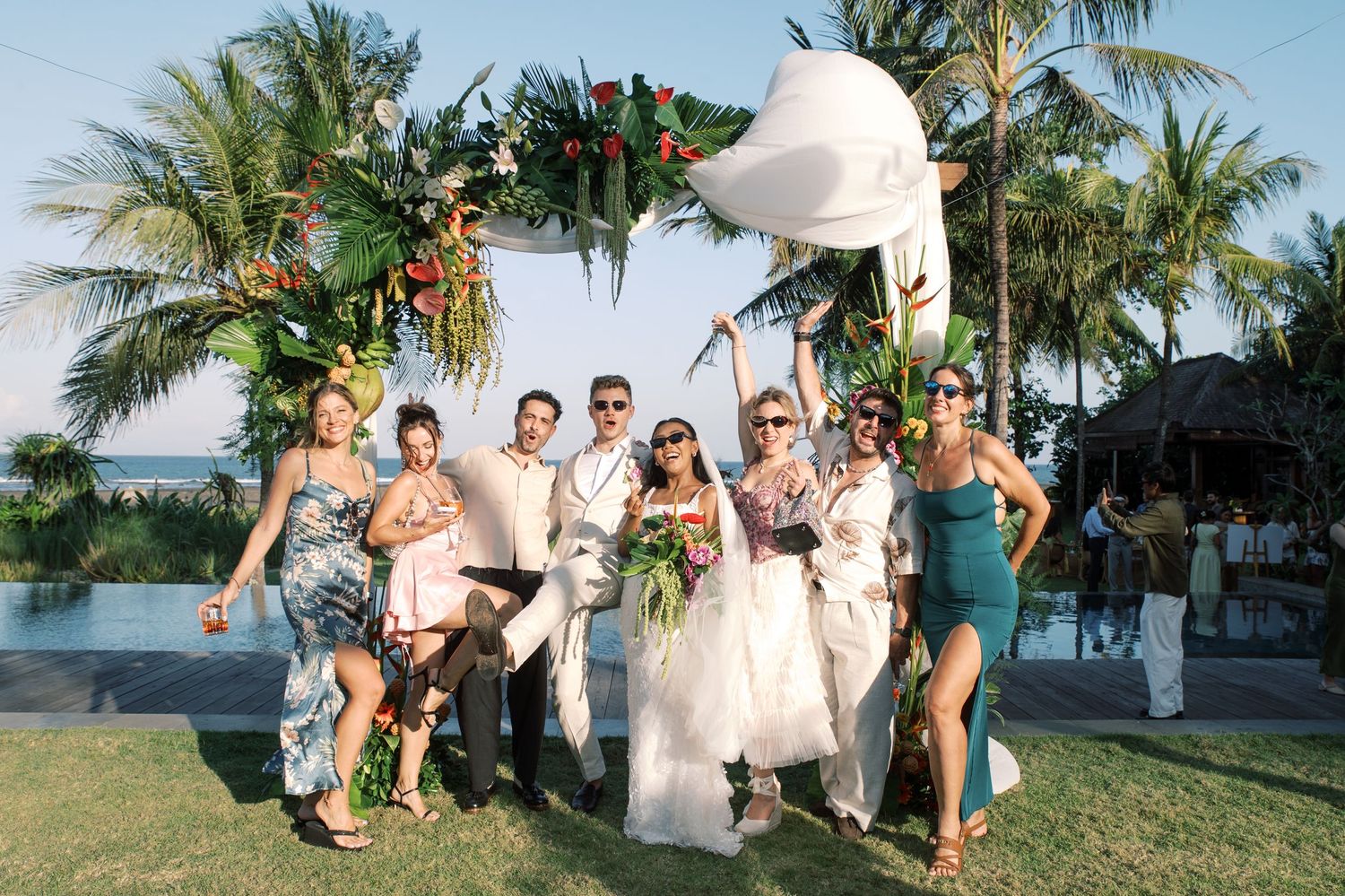 Wedding party poses under tropical floral arch near poolside at beach venue.