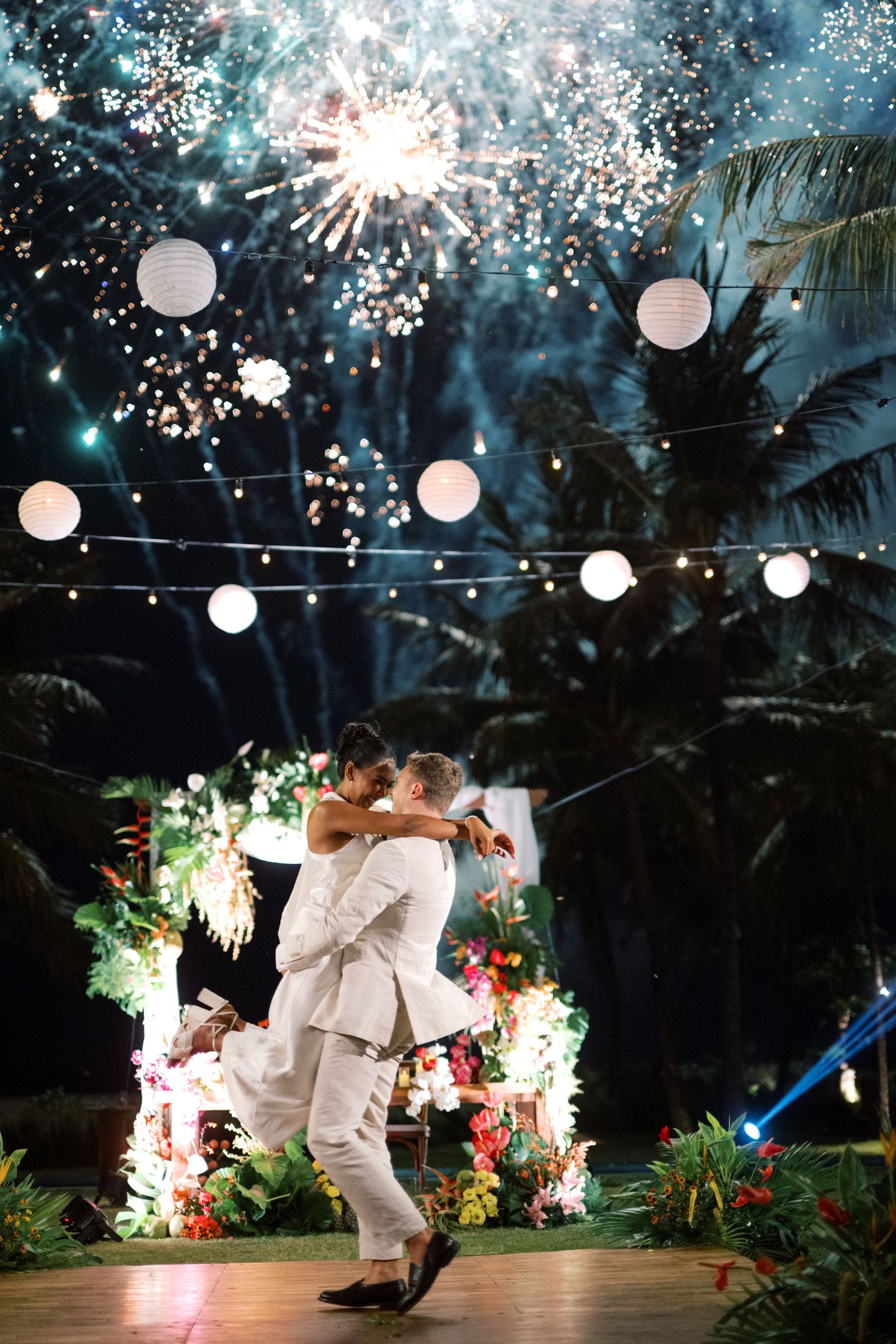 Two grooms share first dance under fireworks and paper lanterns at tropical outdoor wedding celebration at night.