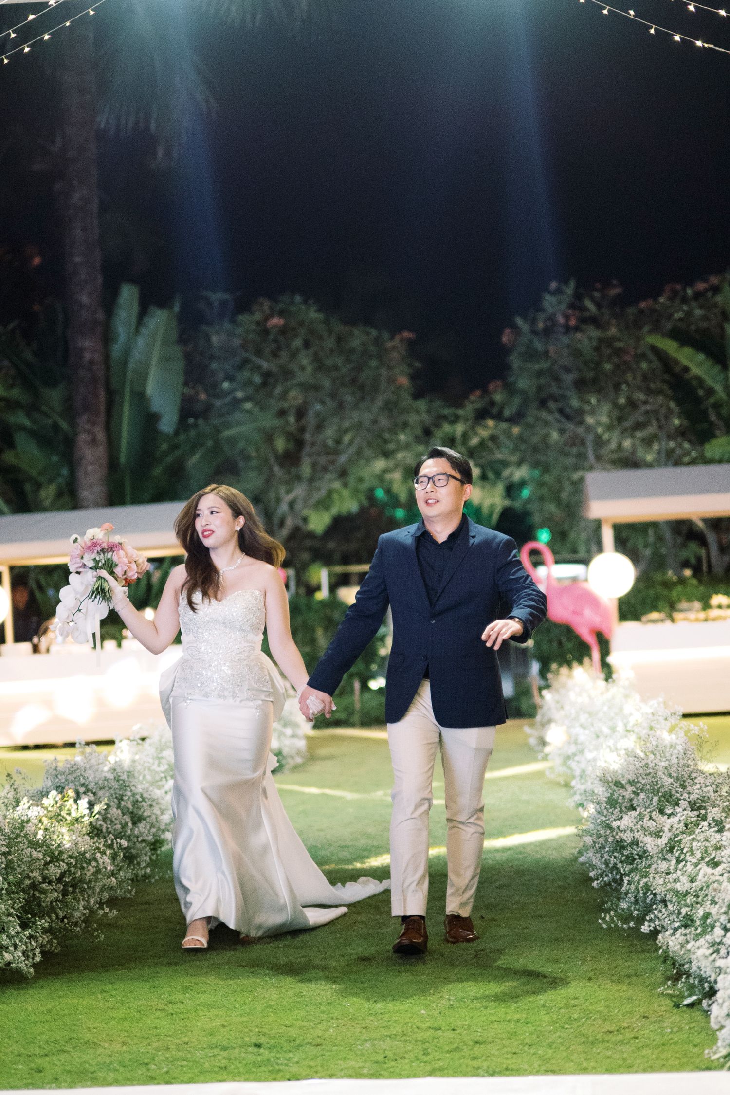 Bride in white gown and groom in navy suit walk hand-in-hand at outdoor evening wedding reception.